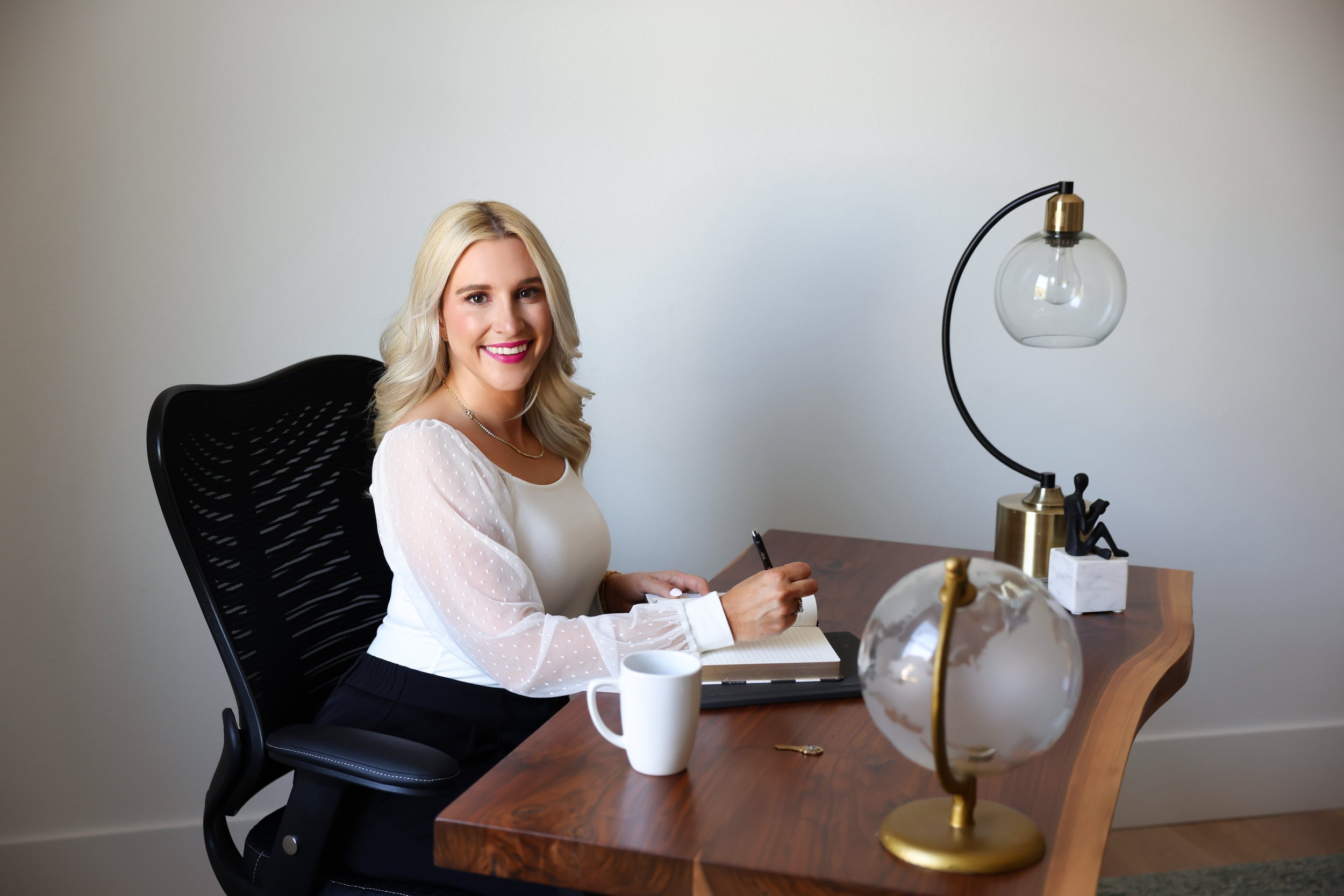 A woman with blonde hair sitting at a desk, smiling, wearing a white blouse with sheer polka dot sleeves, writing in a notebook. The desk has a white mug, a globe, and a modern lamp.