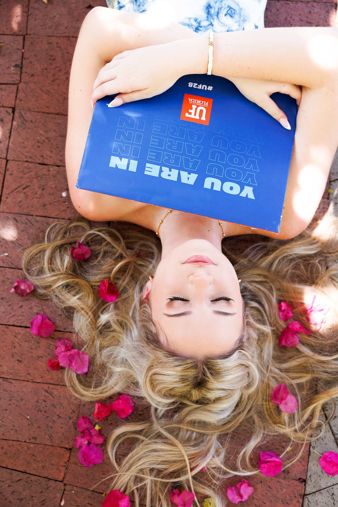 A woman with long blonde curly hair lies on a brick surface with her eyes closed, surrounded by pink and red flower petals, holding a blue book over her chest.
