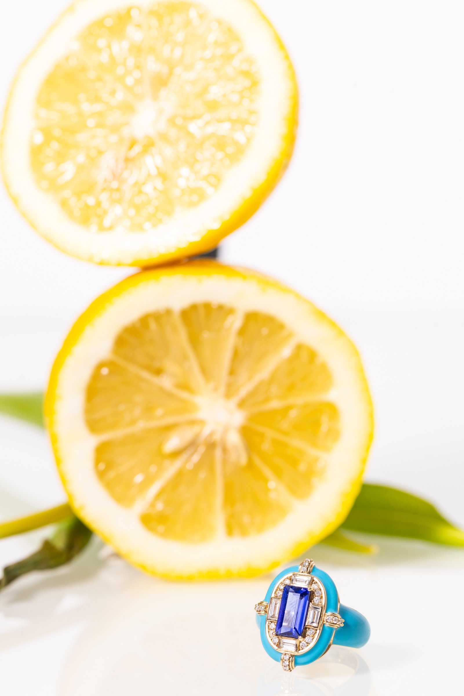 Two lemon halves, one above the other, with a blue ring with a large blue gemstone and smaller diamonds resting on a white surface with green leaves.