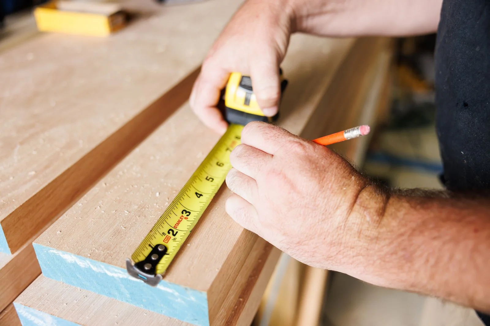 A person measuring a piece of wood with a yellow measuring tape while holding a pencil in their hand.