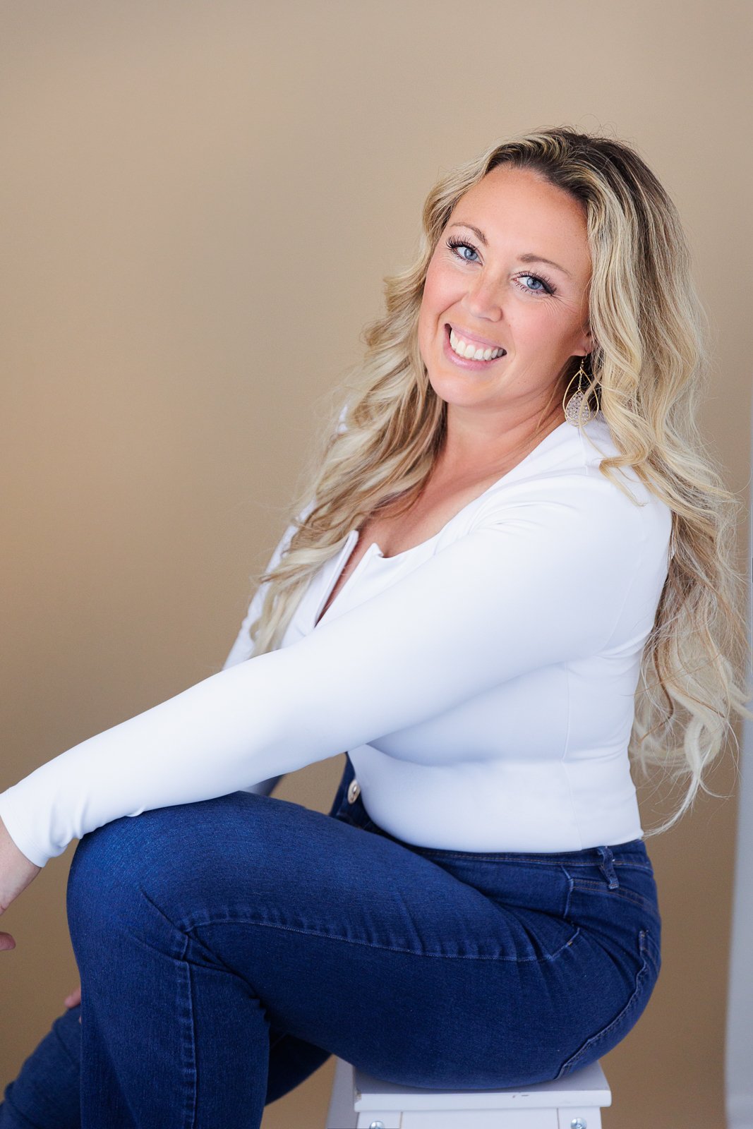 A woman with long, wavy blonde hair, blue eyes, and light skin, smiling and sitting on a stool against a beige background, wearing a white long-sleeve top and dark jeans.