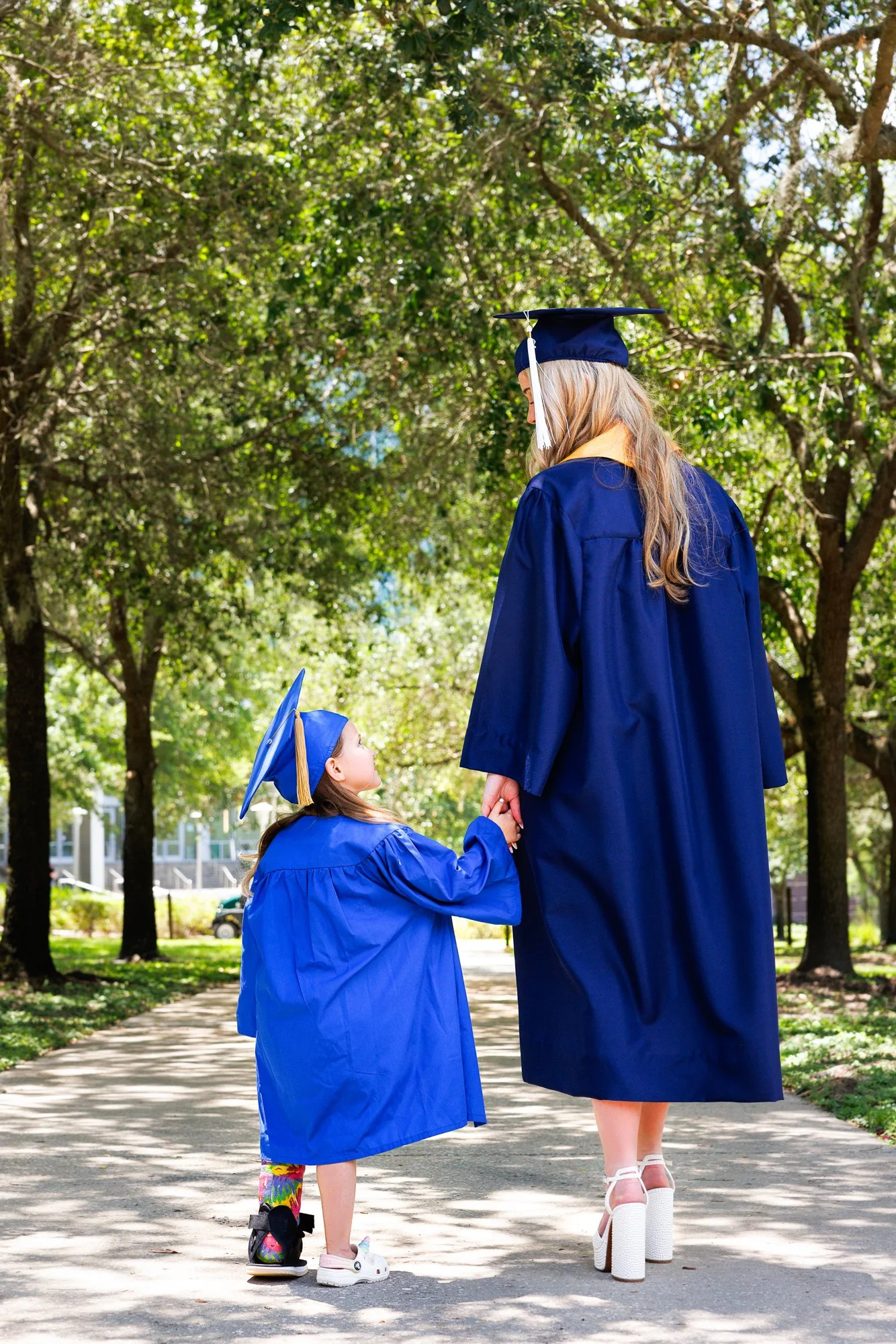 A young girl and woman dressed in blue graduation gowns and caps holding hands outdoors on a sunny day, surrounded by green trees.