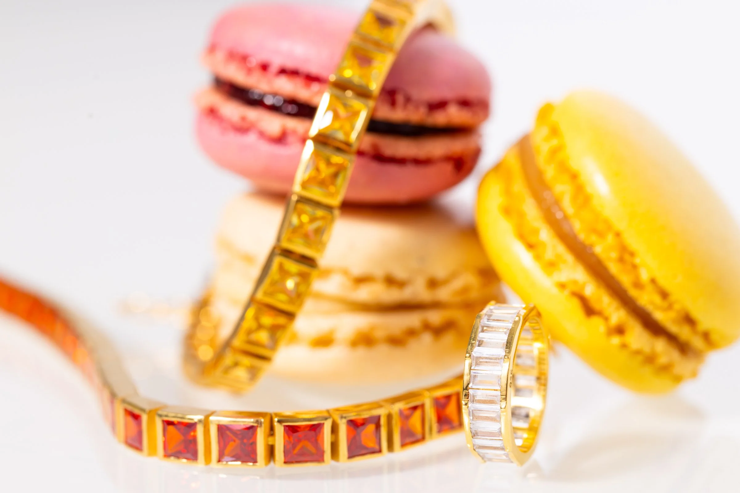 Close-up of three colorful macarons in pink, yellow, and cream, paired with two gold and gemstone rings, on a white background.