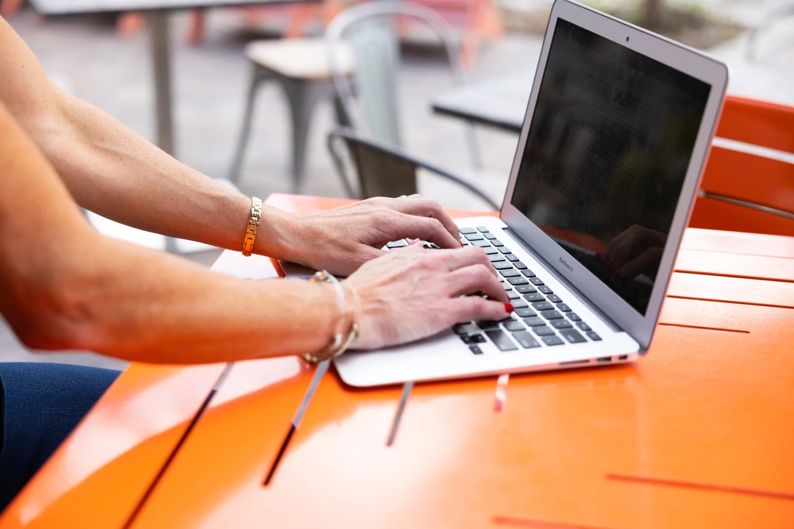 A person typing on a silver MacBook laptop at an orange outdoor table.