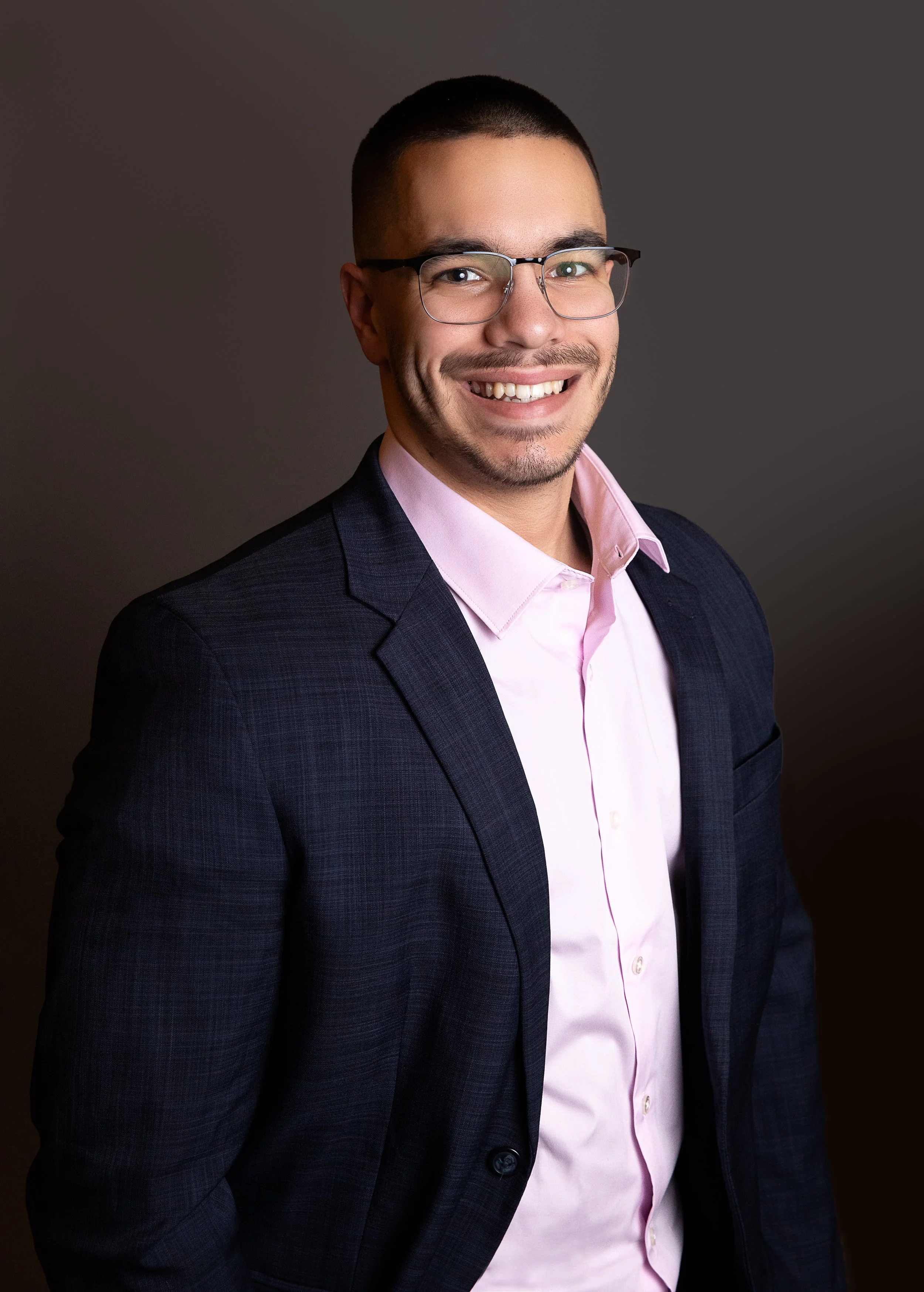 A professional headshot of a man with glasses, short dark hair, wearing a dark suit jacket and a light pink shirt, smiling against a dark gradient background.