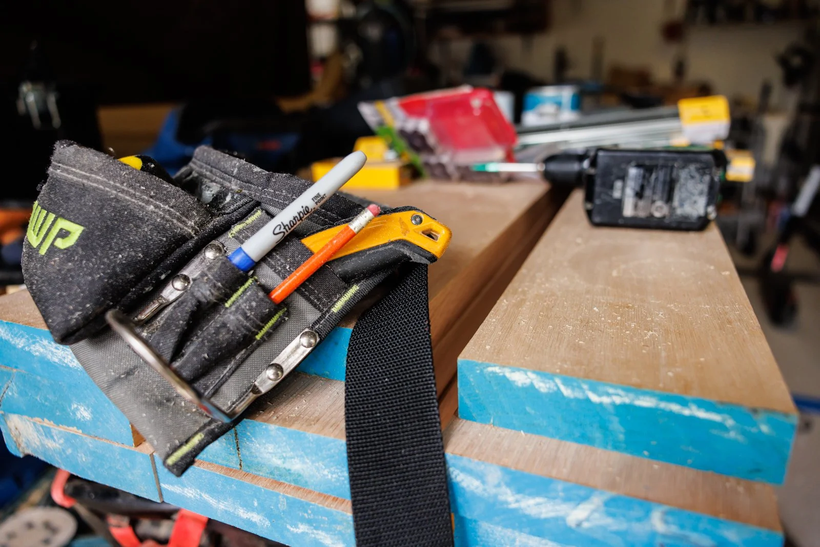 A workbench with tools and materials, including a tool pouch with a Sharpie marker and a pen, in a workshop setting.
