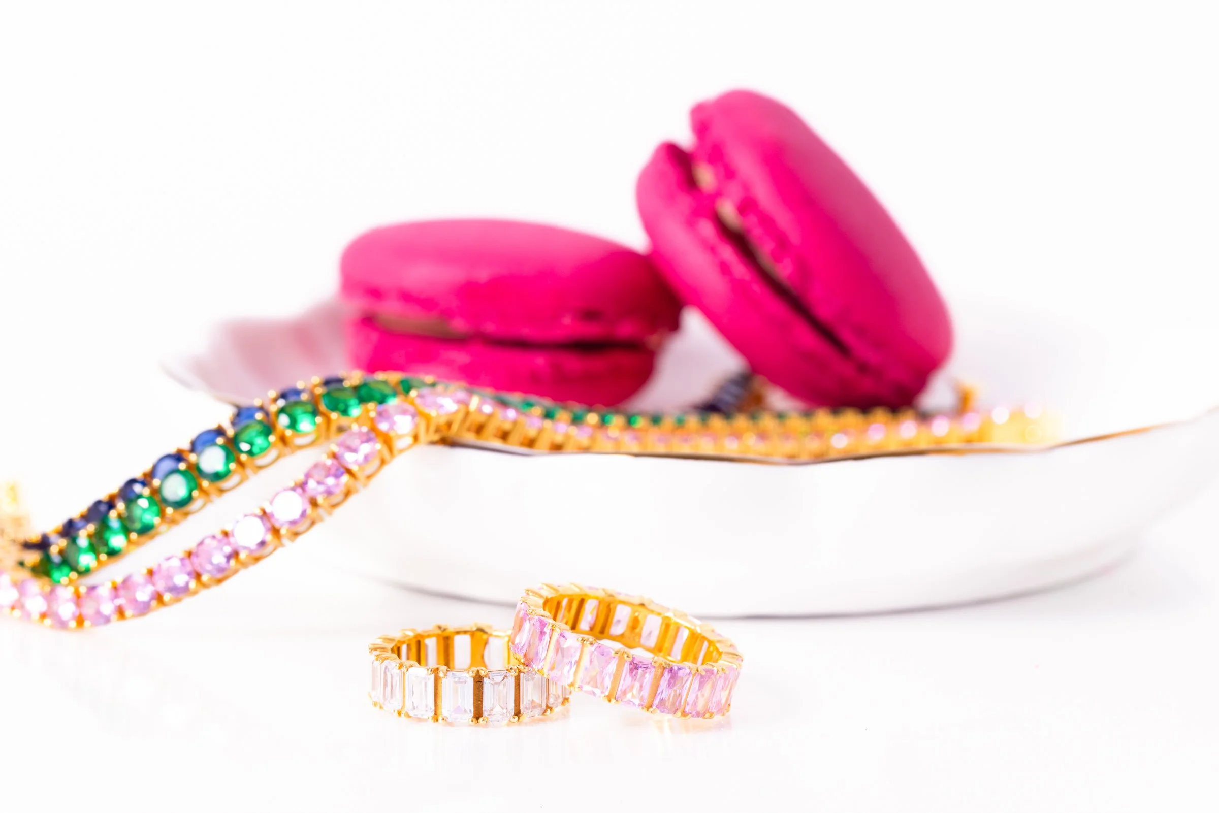 Pink macarons on a white plate with colorful jeweled bracelets and rings in the foreground.