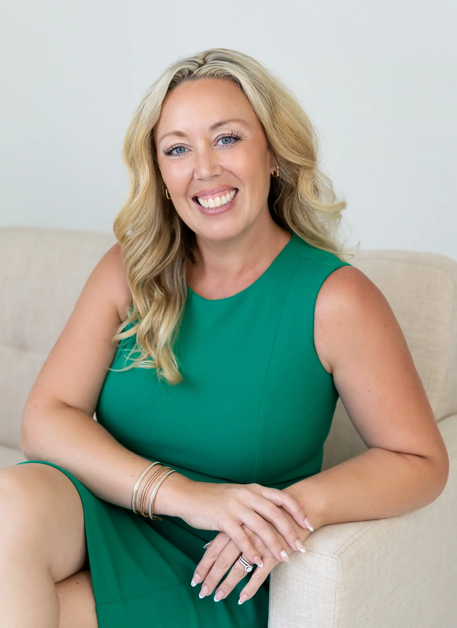 A woman with blonde hair and blue eyes, wearing a green sleeveless dress, sitting on a beige sofa and smiling at the camera.
