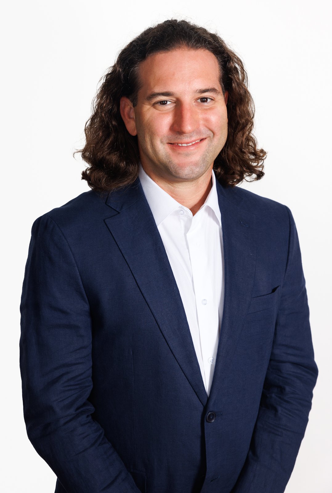 Headshot of a man with long, curly brown hair wearing a navy suit and white dress shirt against a white background.