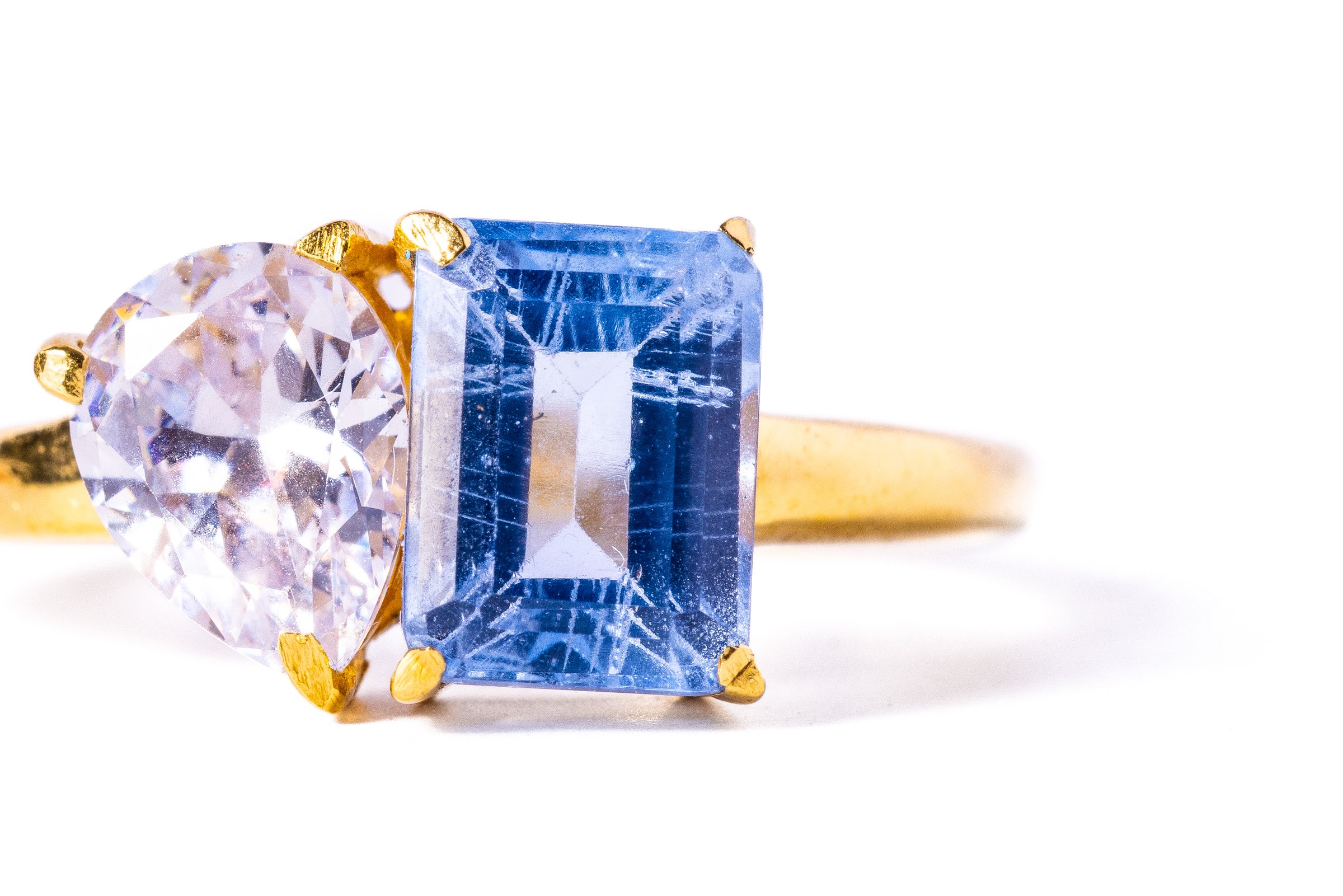 Close-up of a gold ring with a large, clear diamond and a prominent rectangular blue gemstone set in a prong setting. The image is against a white background.