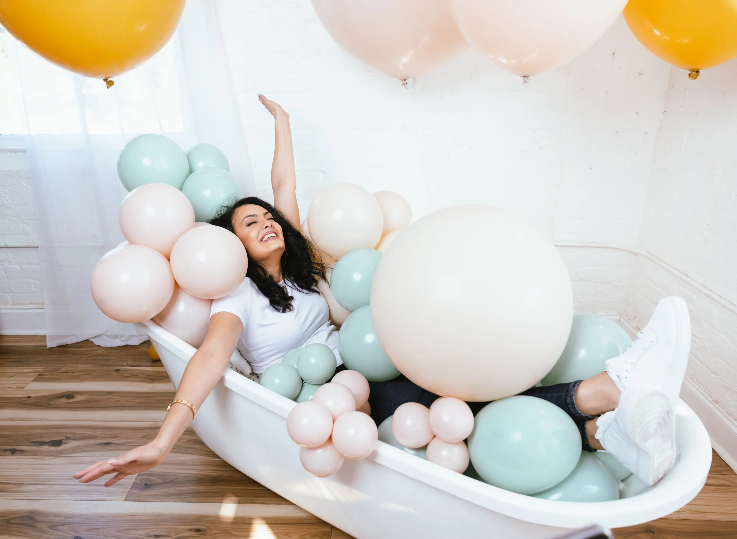 Woman smiling and lying inside a bathtub filled with pastel-colored balloons, with some floating above her.