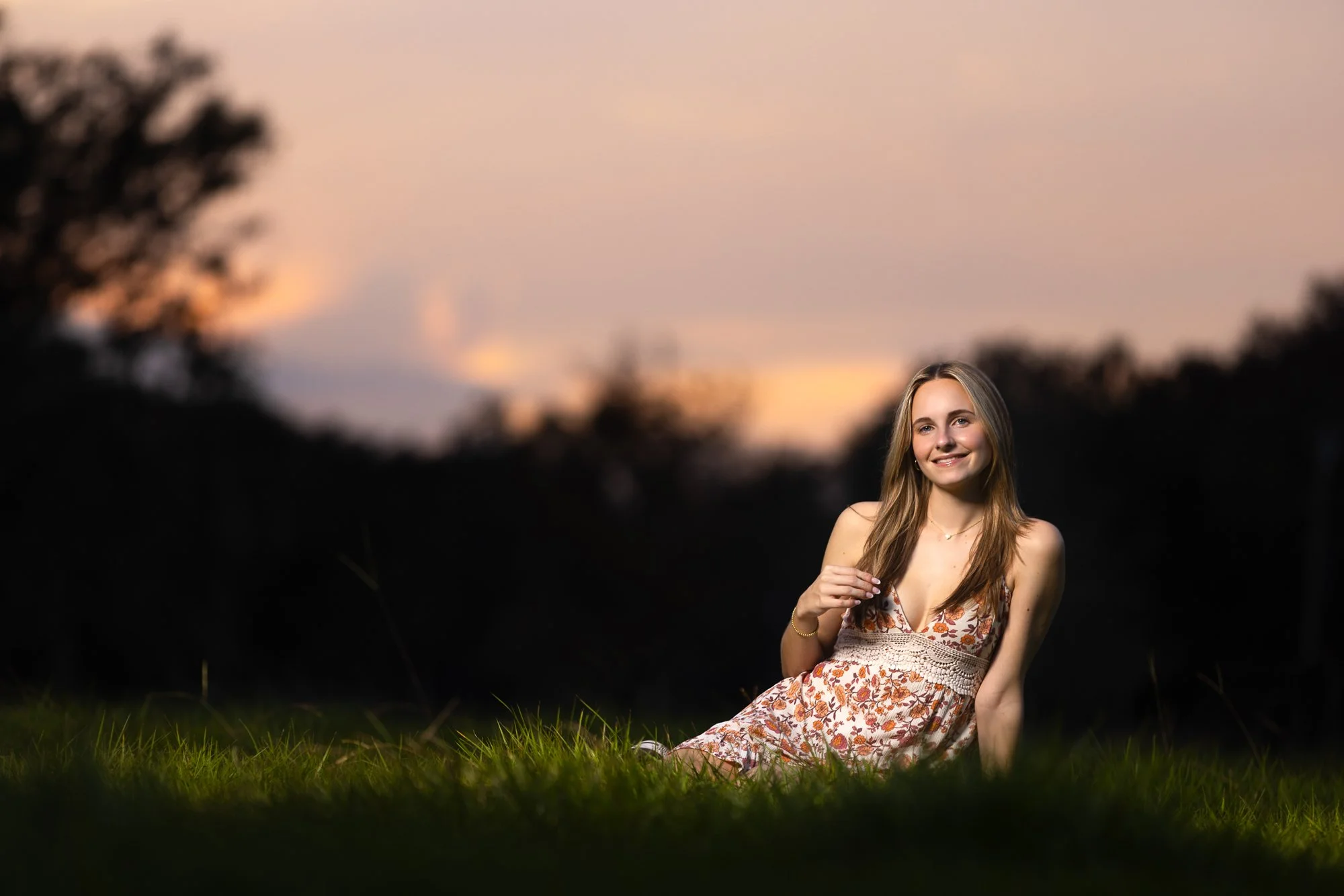A young woman in a floral summer dress sitting on the grass at sunset, smiling and looking at the camera.