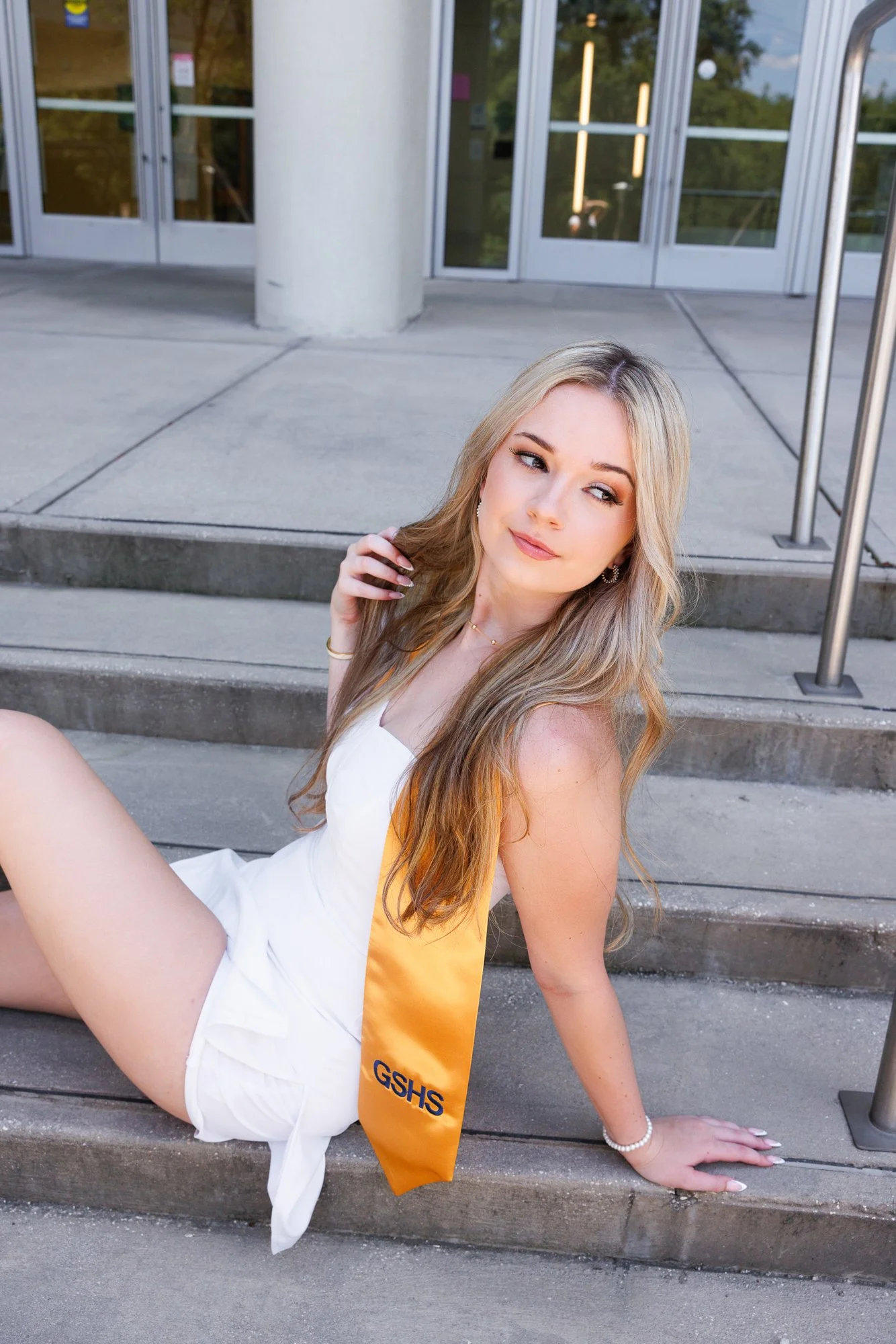 A young woman with long blonde hair sitting on concrete stairs outside a building, wearing a white dress and an orange sash with the letters "GSHS". She has a thoughtful expression and is looking to her right, with a bracelet and earrings visible.
