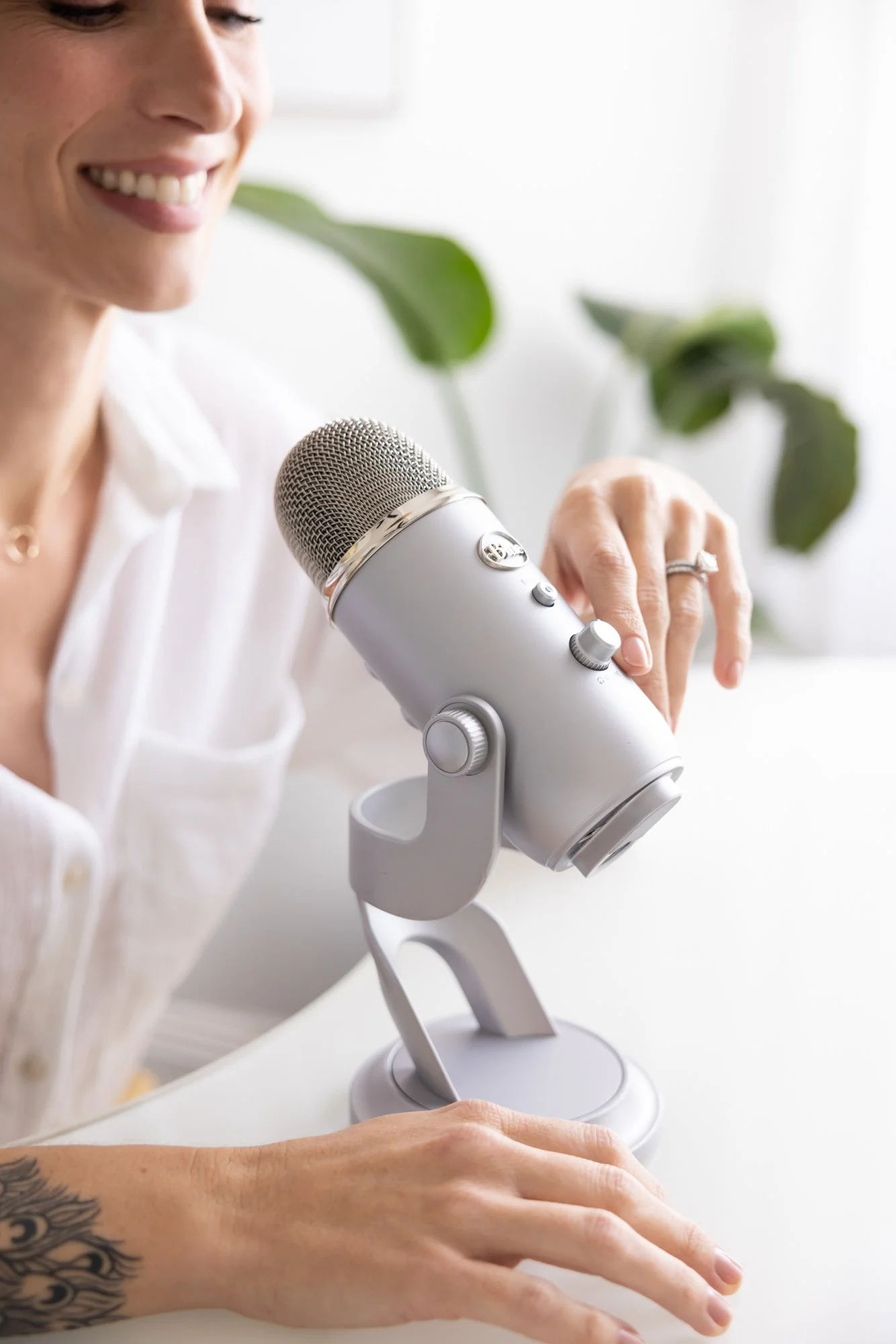 Close-up of a woman with a tattoo on her arm, smiling and speaking into a gray microphone with adjustment knobs, set on a stand. She is wearing a ring and a white shirt, with plants in the background.