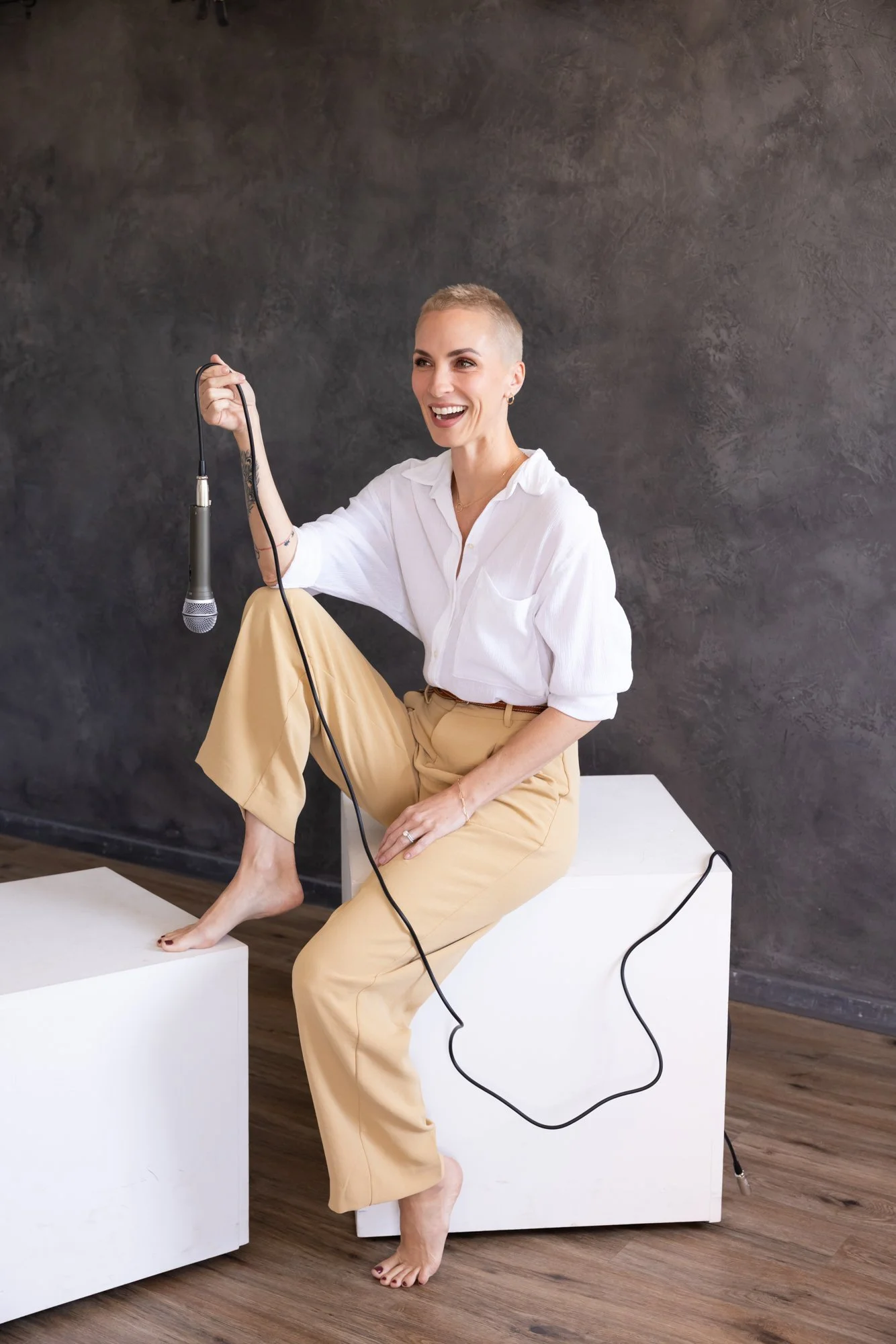 A woman with short hair, wearing a white blouse and beige pants, sitting barefoot on a white block, holding a microphone, smiling, with a dark textured wall in the background.