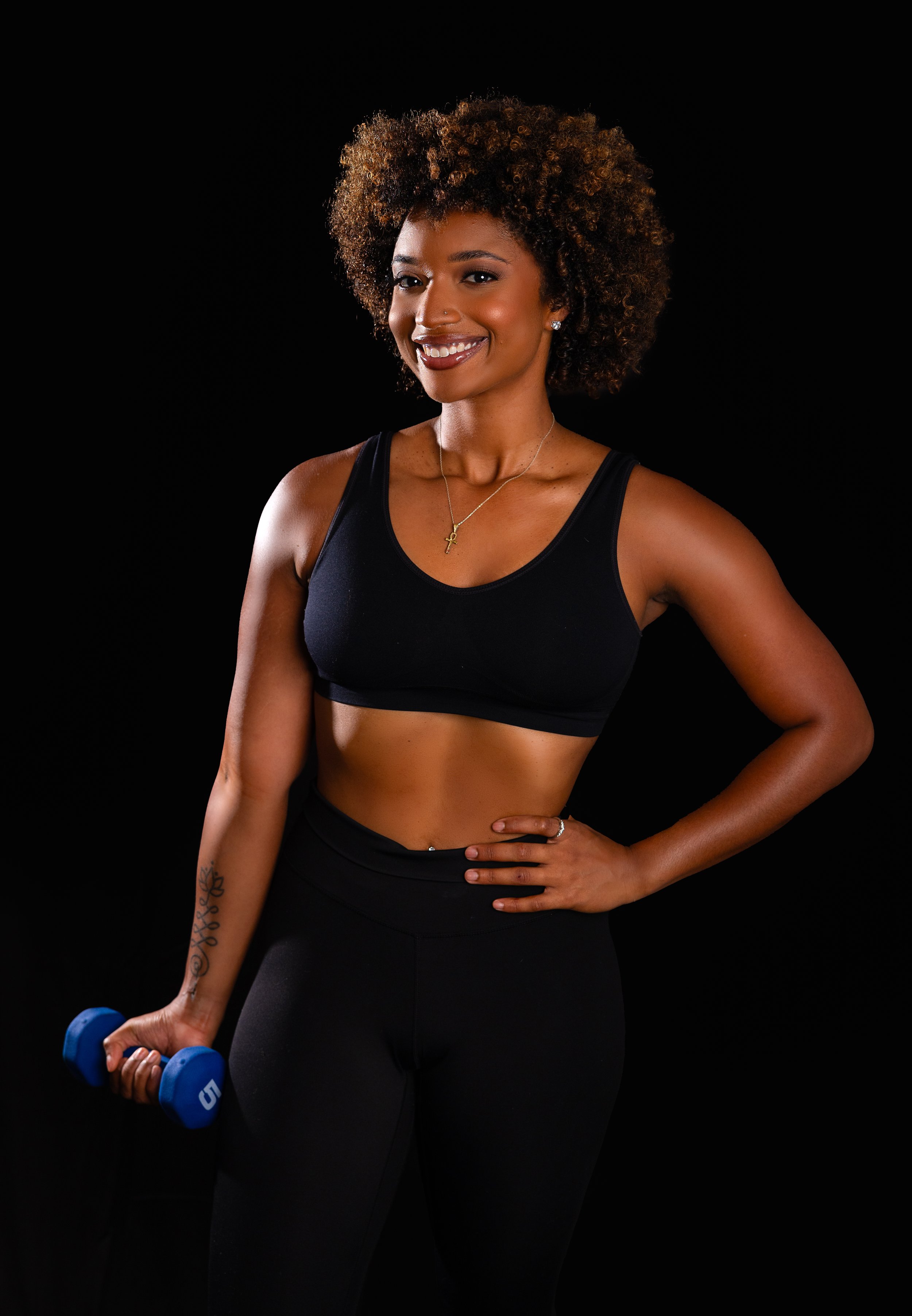 A young woman with curly hair wearing a black sports bra and black leggings, holding a blue dumbbell in her right hand, standing against a black background, smiling at the camera.