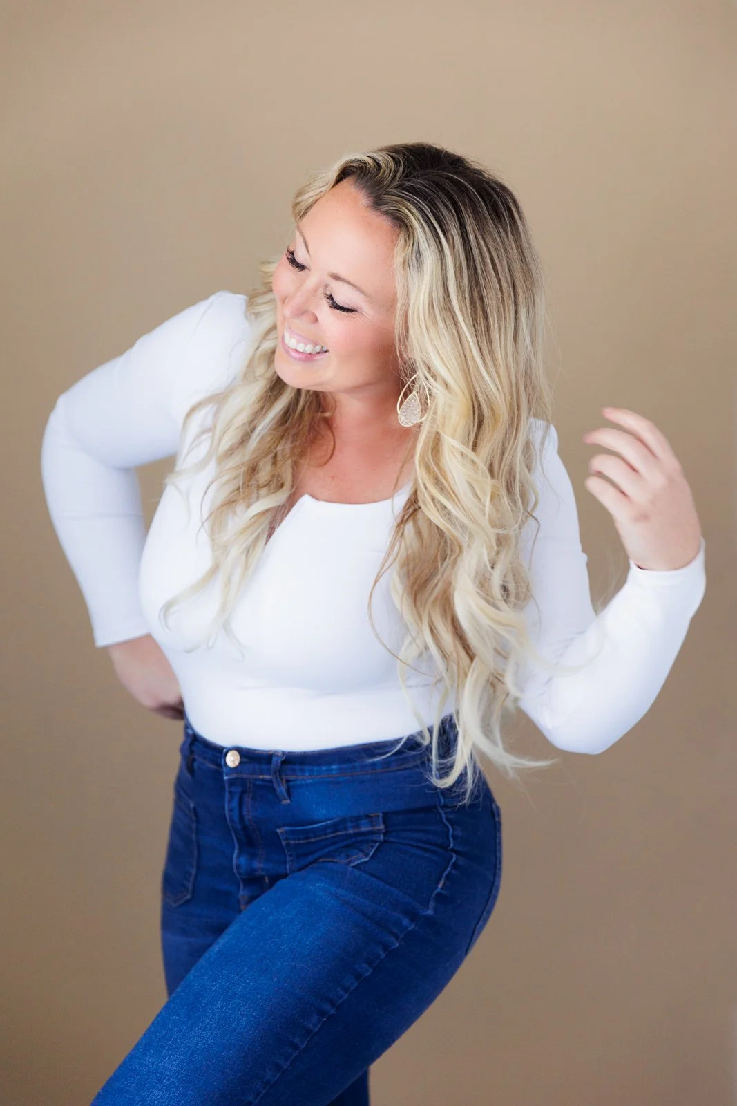 A woman with long, wavy blonde hair in a white top and blue jeans, smiling and looking down, against a plain beige background.