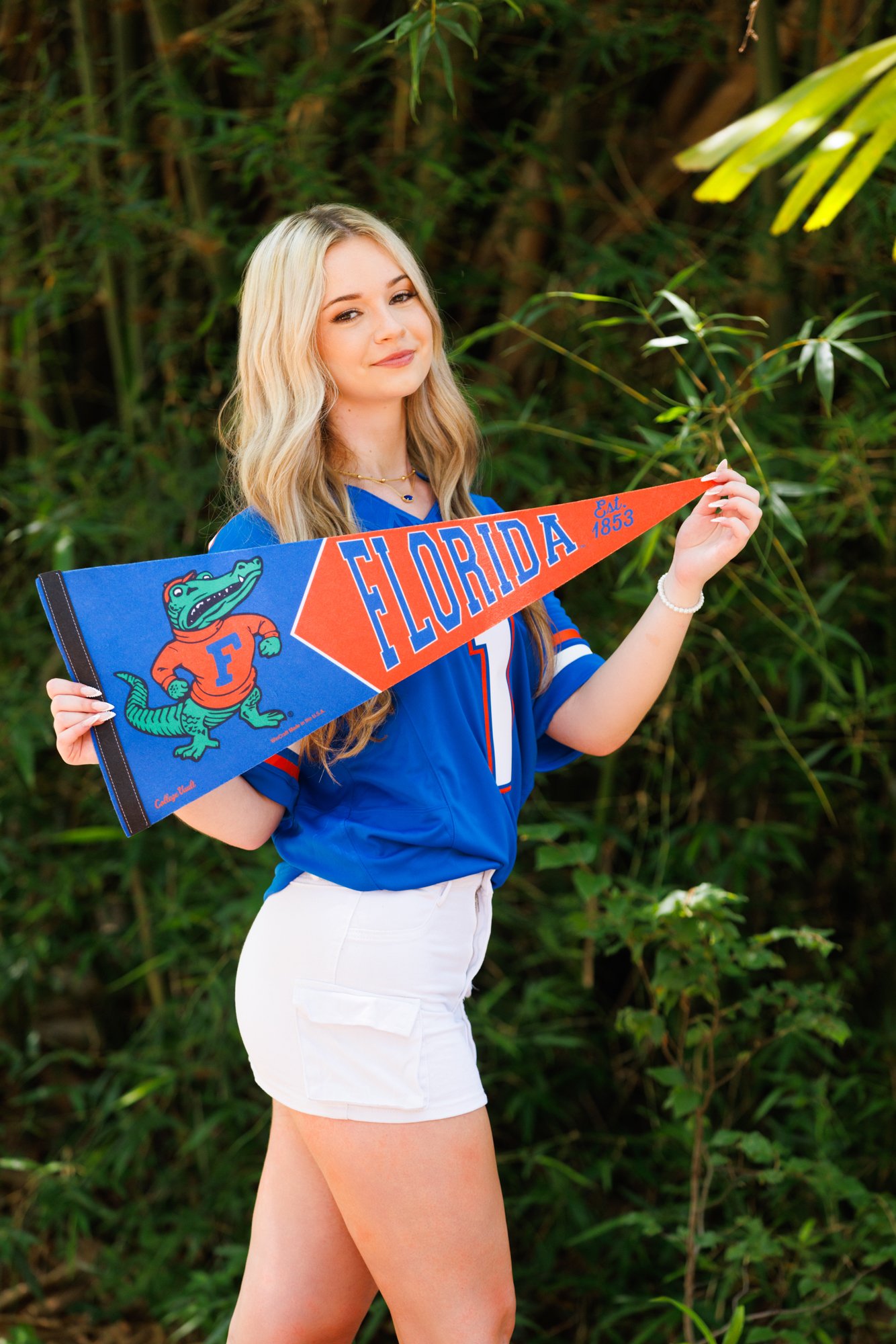 A young woman with long blonde hair is standing outdoors in front of dense green foliage, holding a Florida Gators pennant celebrating the university, smiling slightly.