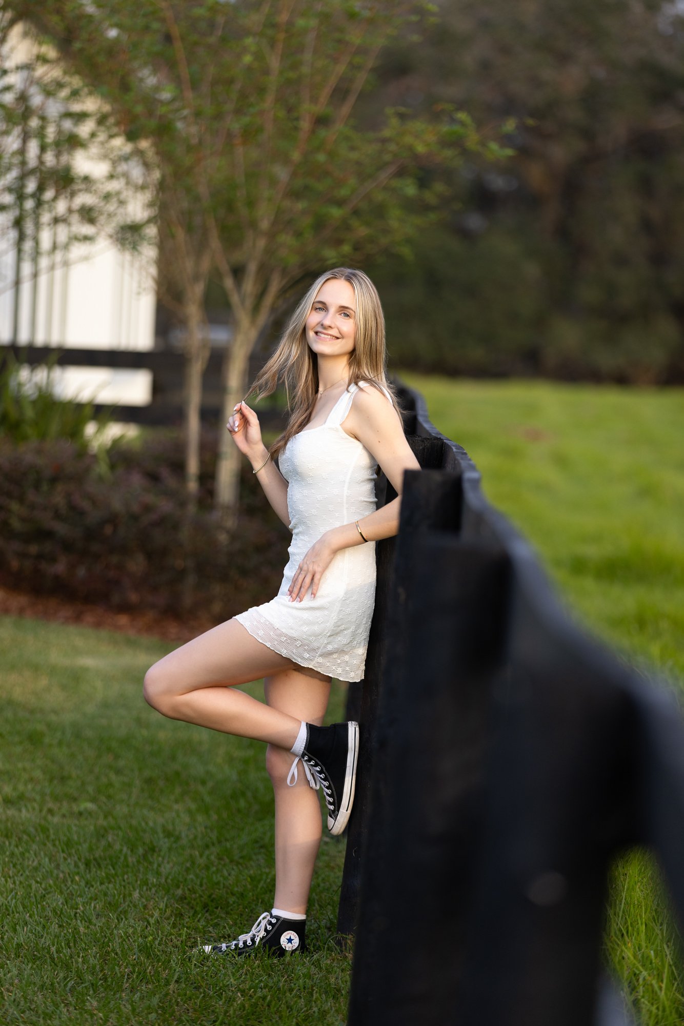 A young woman with long blonde hair in a white dress and black sneakers stands outside near a black fence, smiling at the camera with a lush yard and trees in the background.