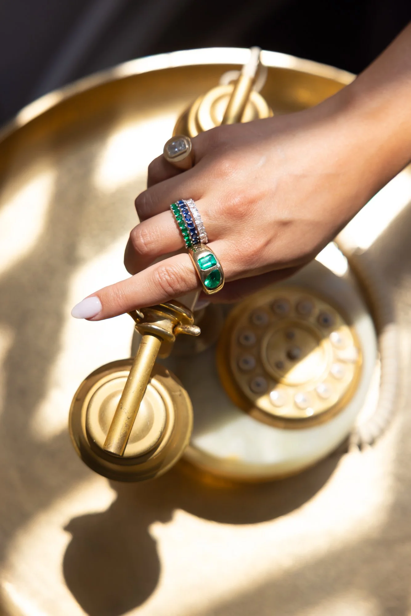 A woman's hand with multiple rings, including one with an emerald, holding a vintage gold telephone receiver.