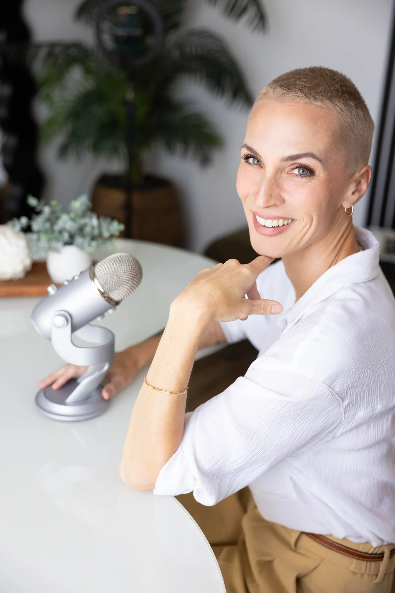 Woman with short blonde hair sitting at a white table with a silver microphone, smiling, and touching her neck, in a bright room with plants and flowers in the background.