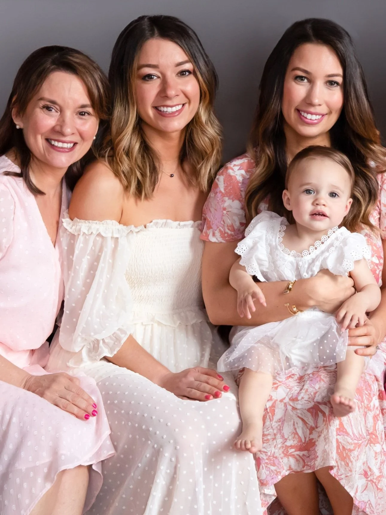Four women, three with dark hair and one with light brown hair, sitting together wearing light-colored, feminine dresses. One woman is holding a young girl dressed in white. All are smiling against a dark background.