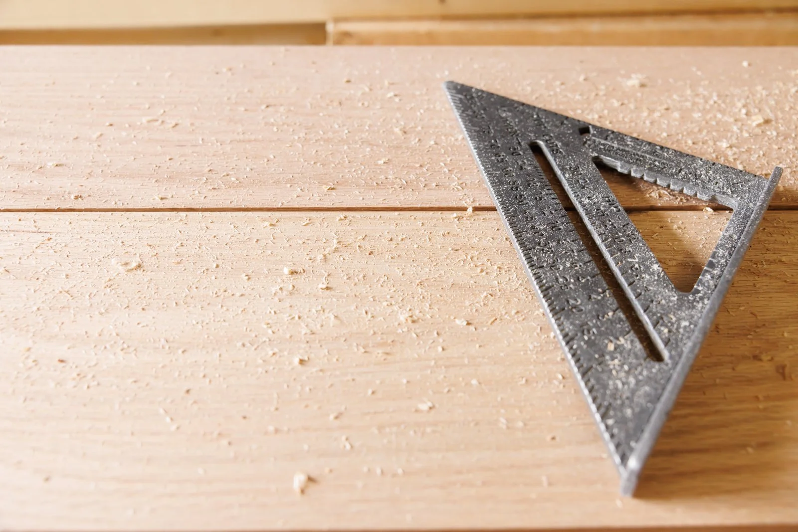A metal carpenter's square lying on a wooden work surface, with wood shavings scattered around.