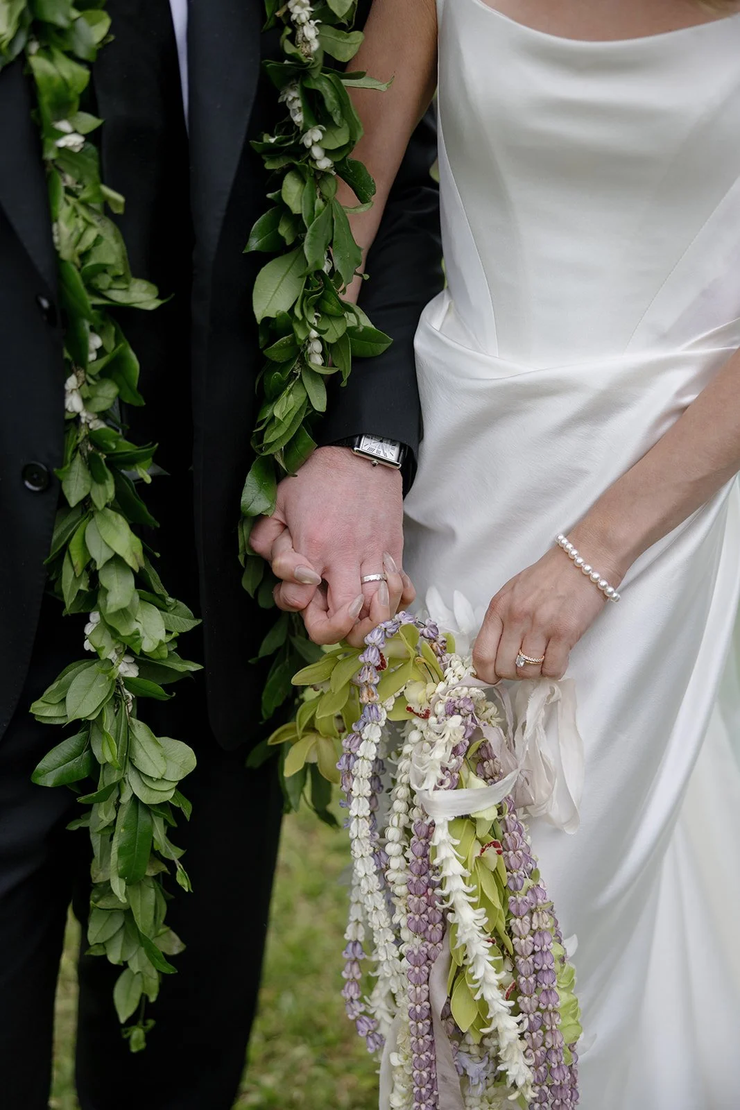Detail shot of bride and groom holding hands at Paliku Gardens wedding, showing groom's lei, bridal bouquet, rings, and dress details