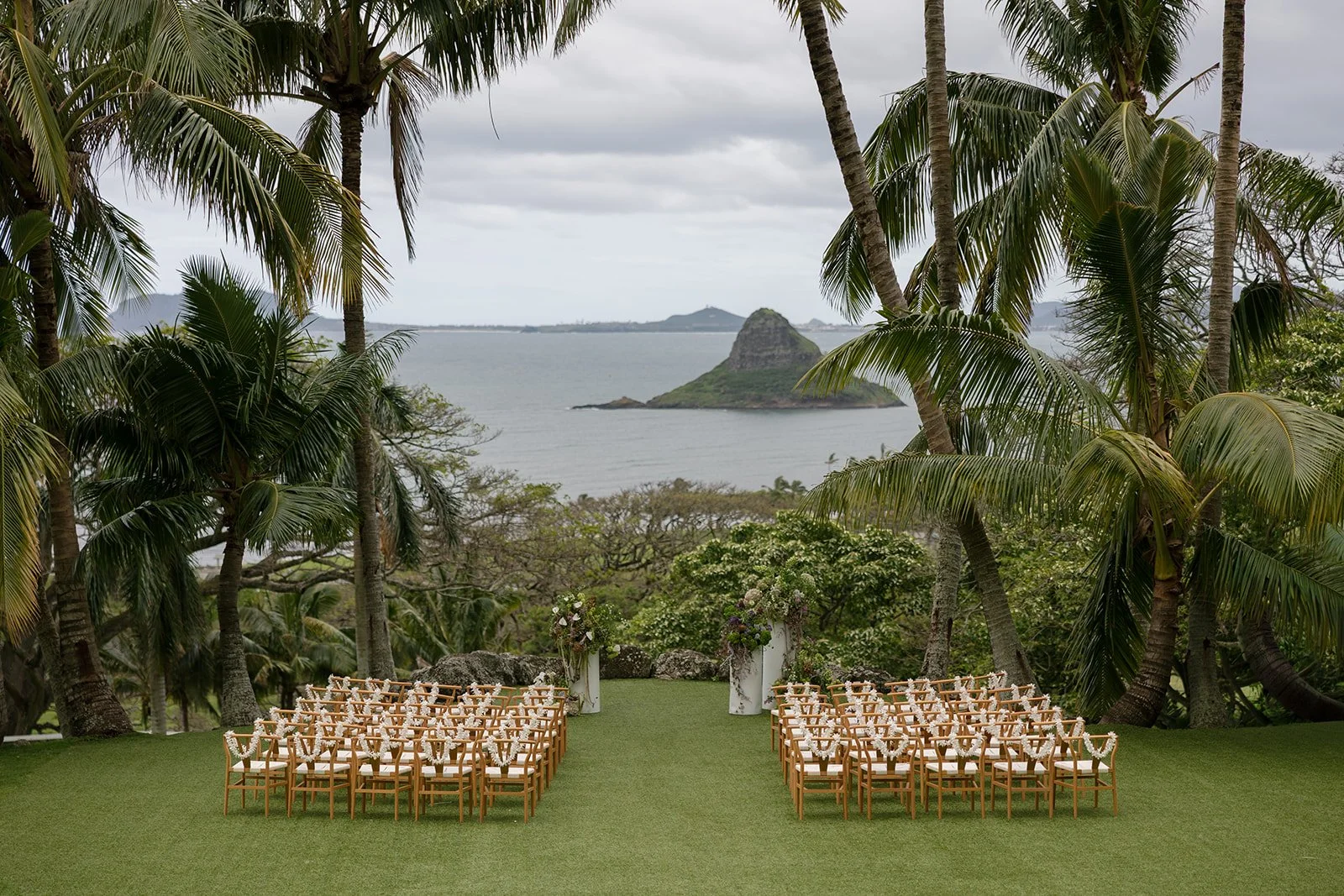 Ceremony at Paliku Gardens with Chinaman's Hat in the background, wishbone chairs with white orchid lei, and floral altar arrangements on white pillars