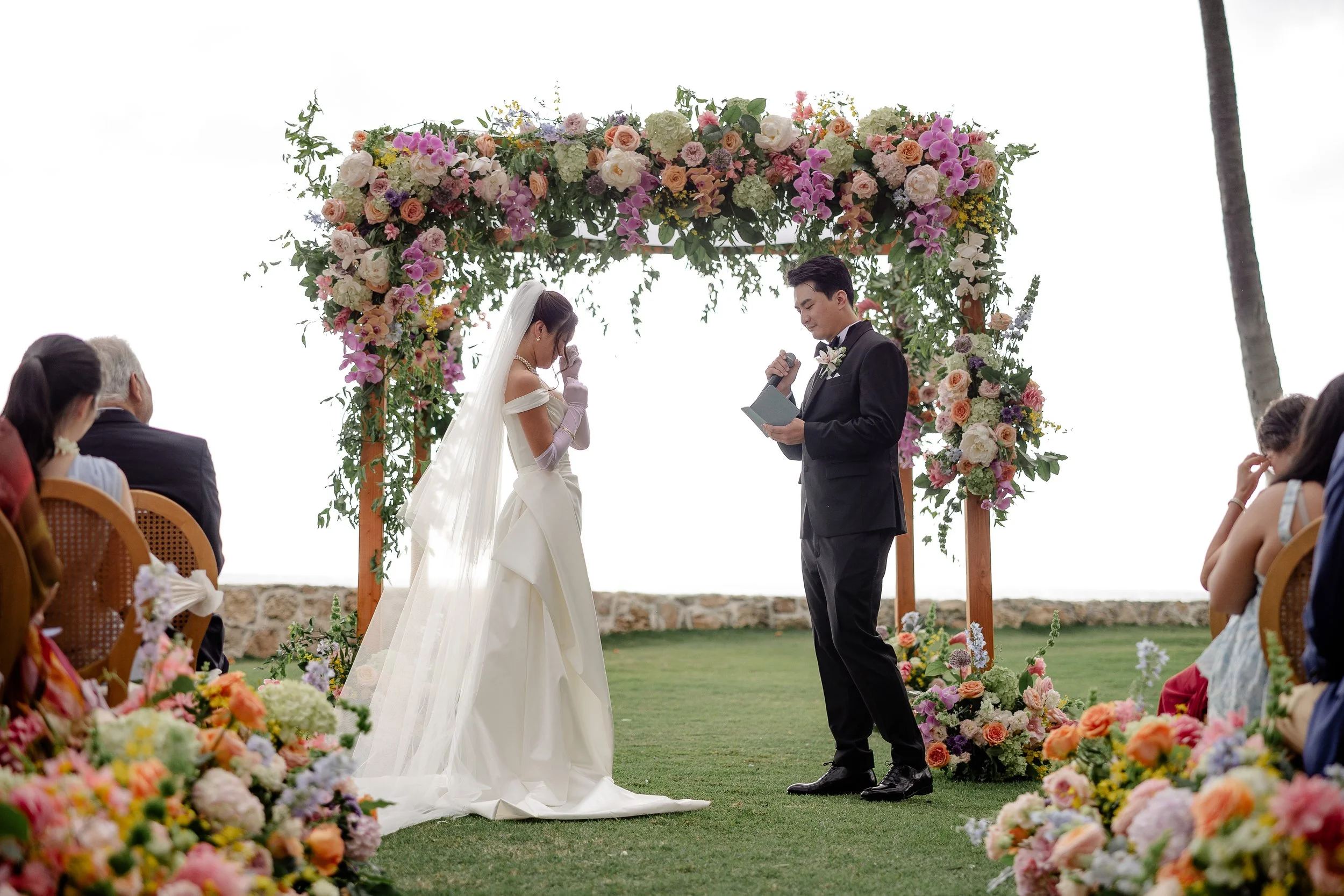 groom exchanging wedding vows while bride is whipping tears from her face, and a wooden alter with cascading flowers is behind them