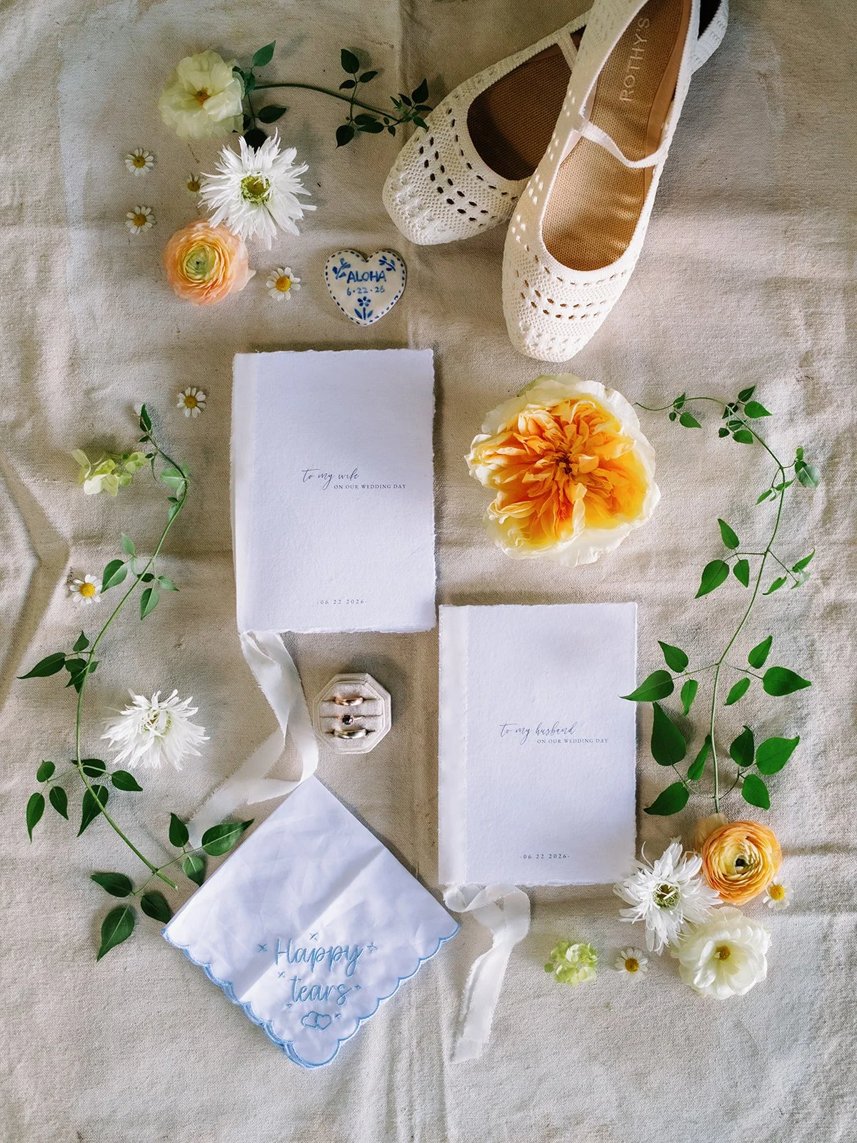 Bride and groom items photographed with a backdrop with vow books, the brides shoes, a handkerchief, and weddings rings with flower styled around the items