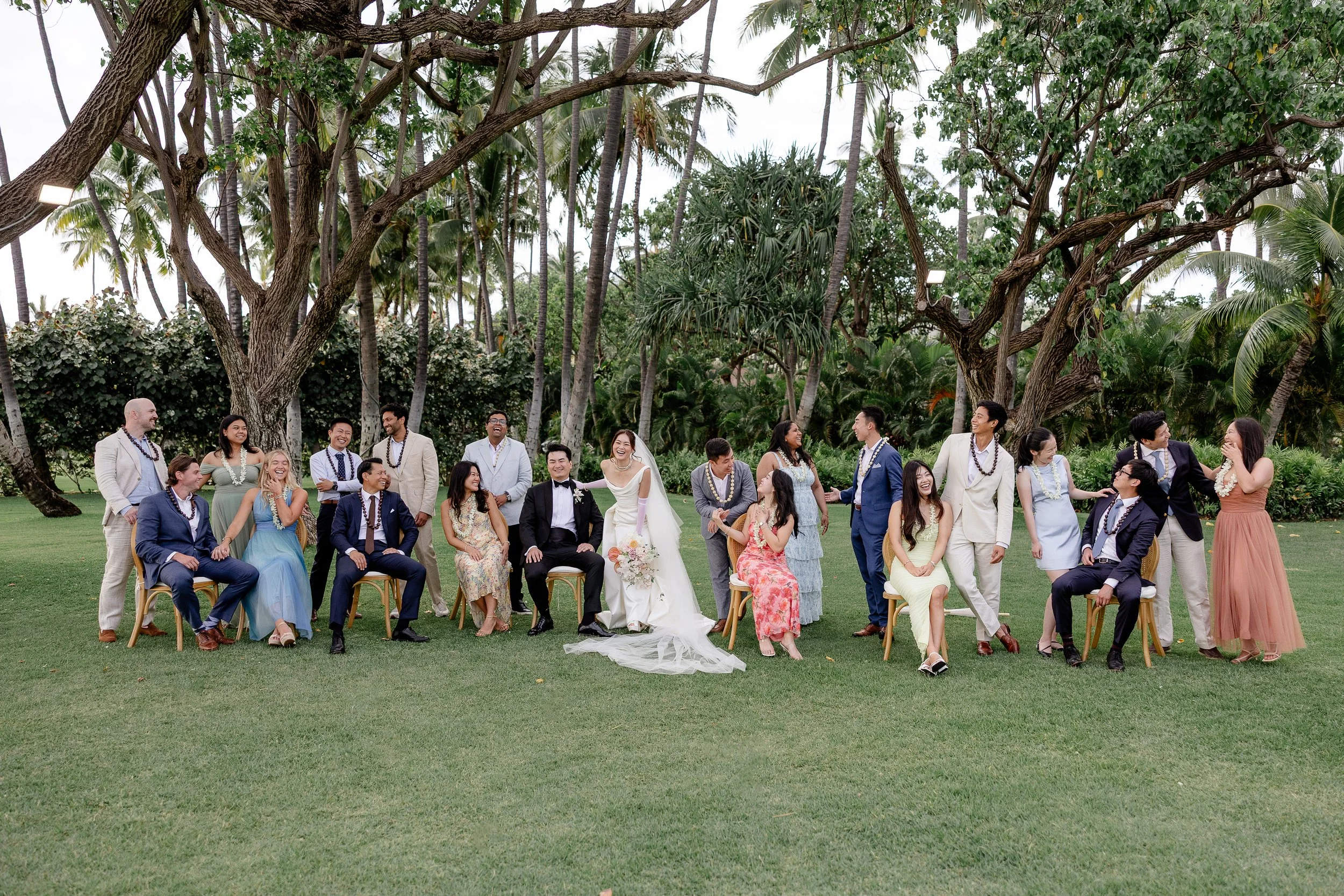 group photo with bride and groom with friends in chairs and standing in a row all smiling and laughing with one another