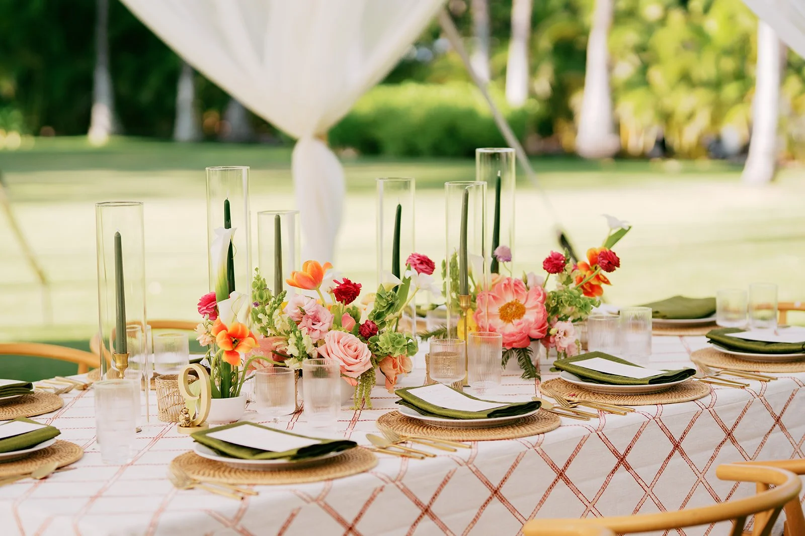 A bride and groom kissing outdoors on a gravel path surrounded by green trees, with the bride holding a bouquet of flowers and wearing a white wedding gown and veil, and the groom in a black suit with a leafy lei. In the lower section, a table is decorated with flowers, candles, glassware, utensils, and a menu card.