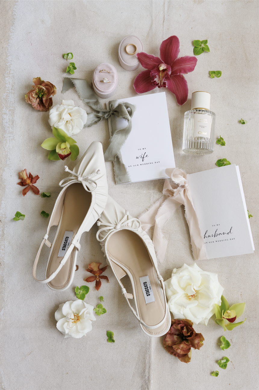 bride and groom detail shot of vow books, wedding rings, perfume, bride's shoes, and scattered flowers