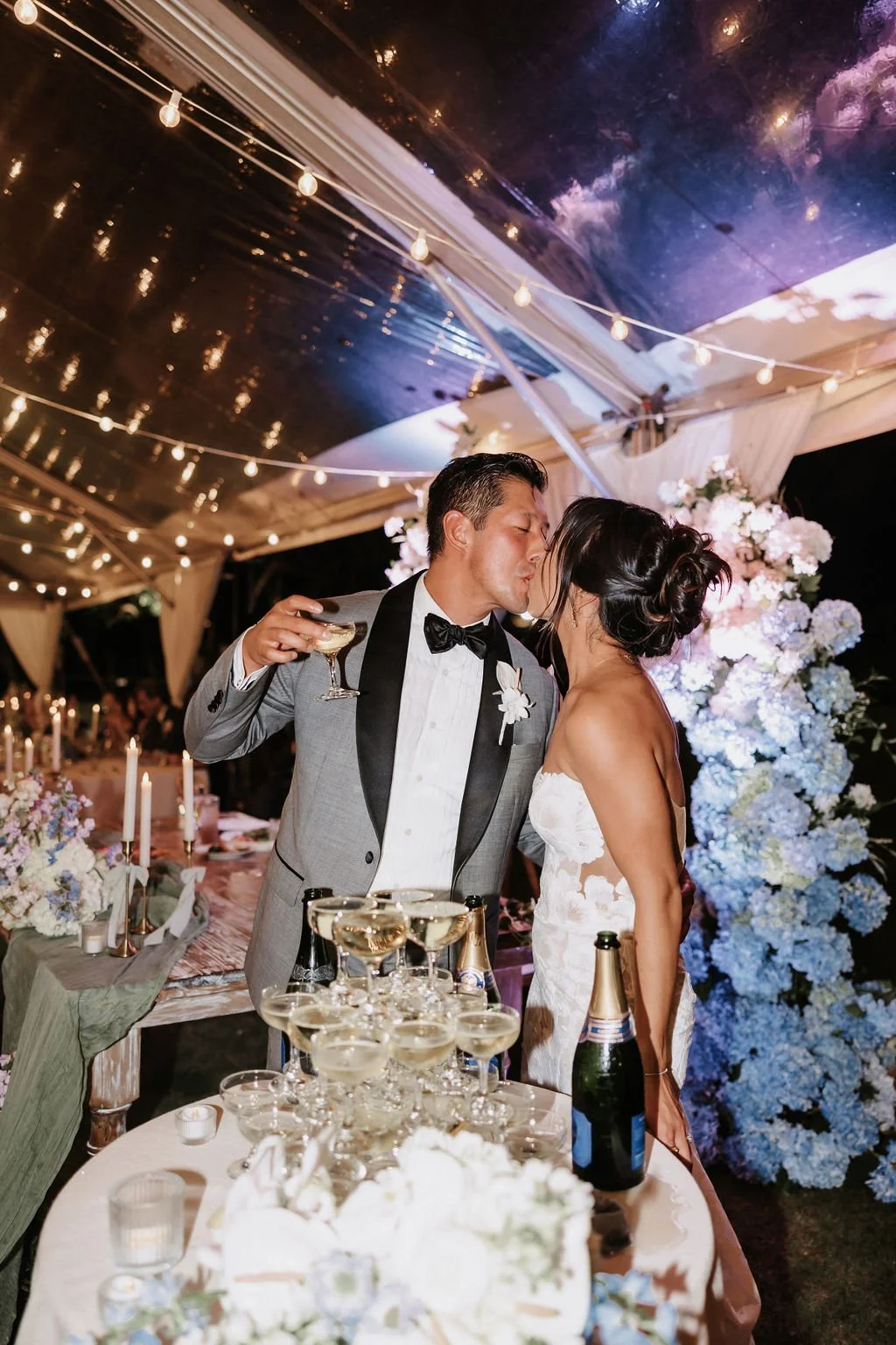 Bride and groom share a kiss while celebrating with a champagne tower during wedding reception.