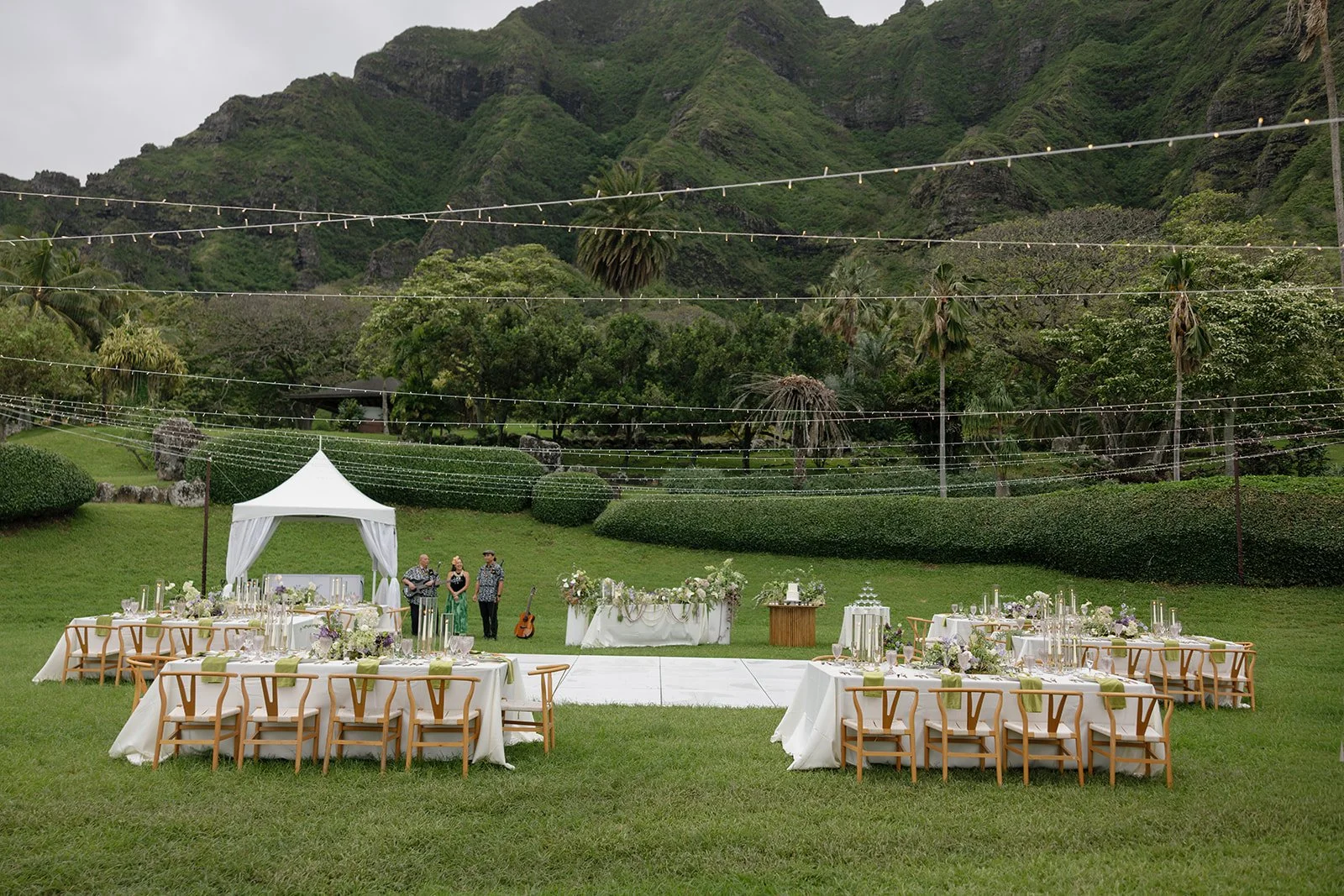 Wide shot of Hawaii wedding reception with twinkle lights, long guest tables, sweetheart table, white dance floor, and Ko'olau mountains in the background at Paliku Gardens