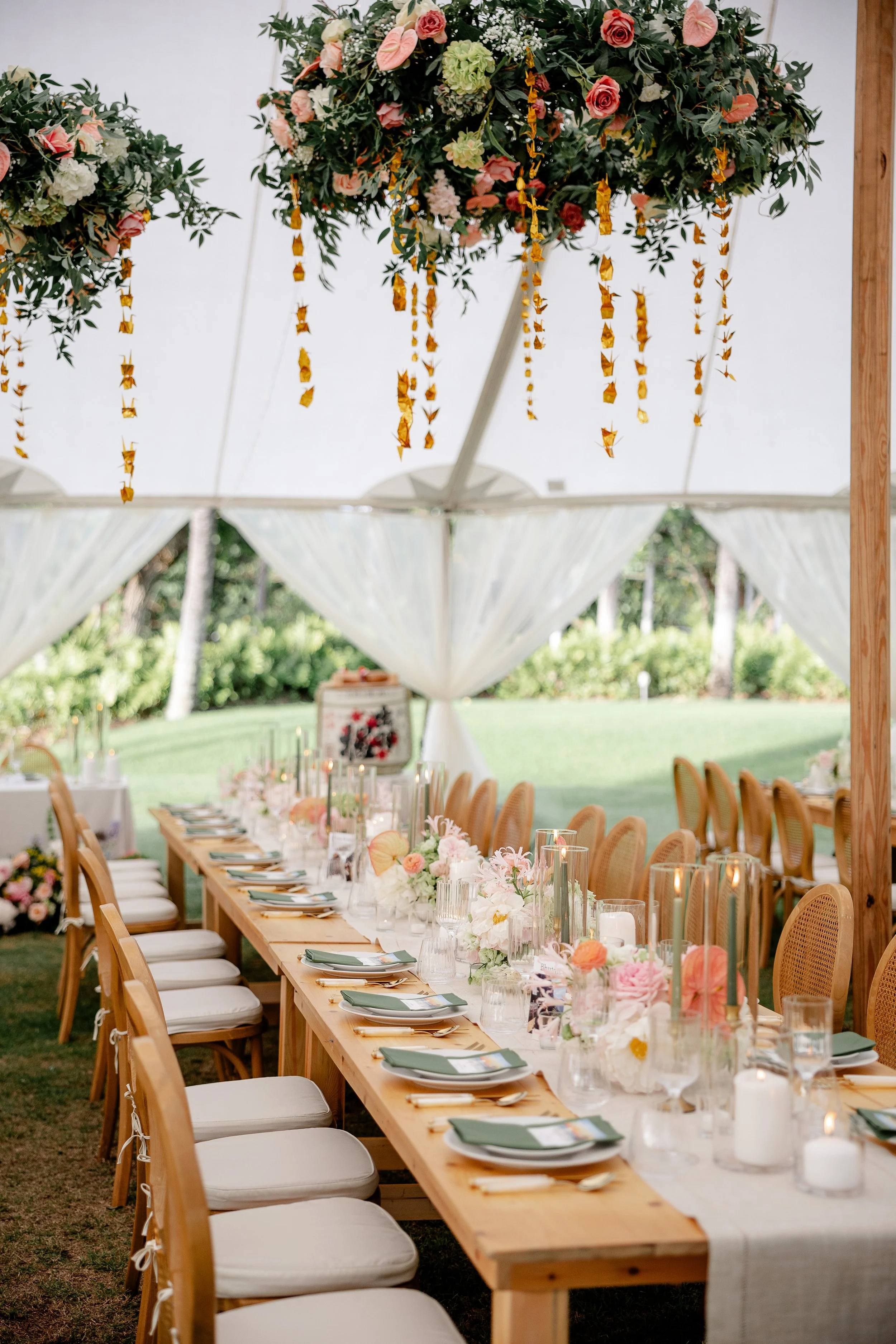 rectangle dining tables with wooden chairs and a cushion. Tables have exposed wood with oatmeal runners, candles, and flower arrangements down the middle. Dinner plates with menus