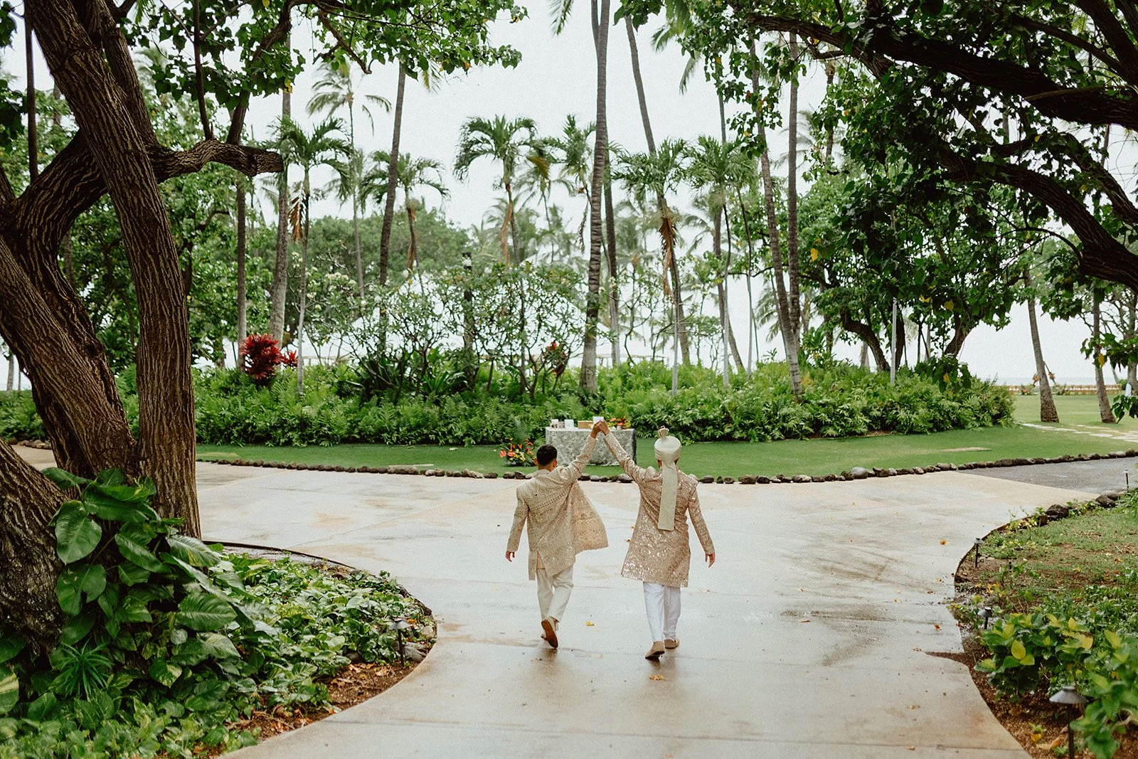 couple holding hands as they walk down the side walk as big trees and greenery surround them