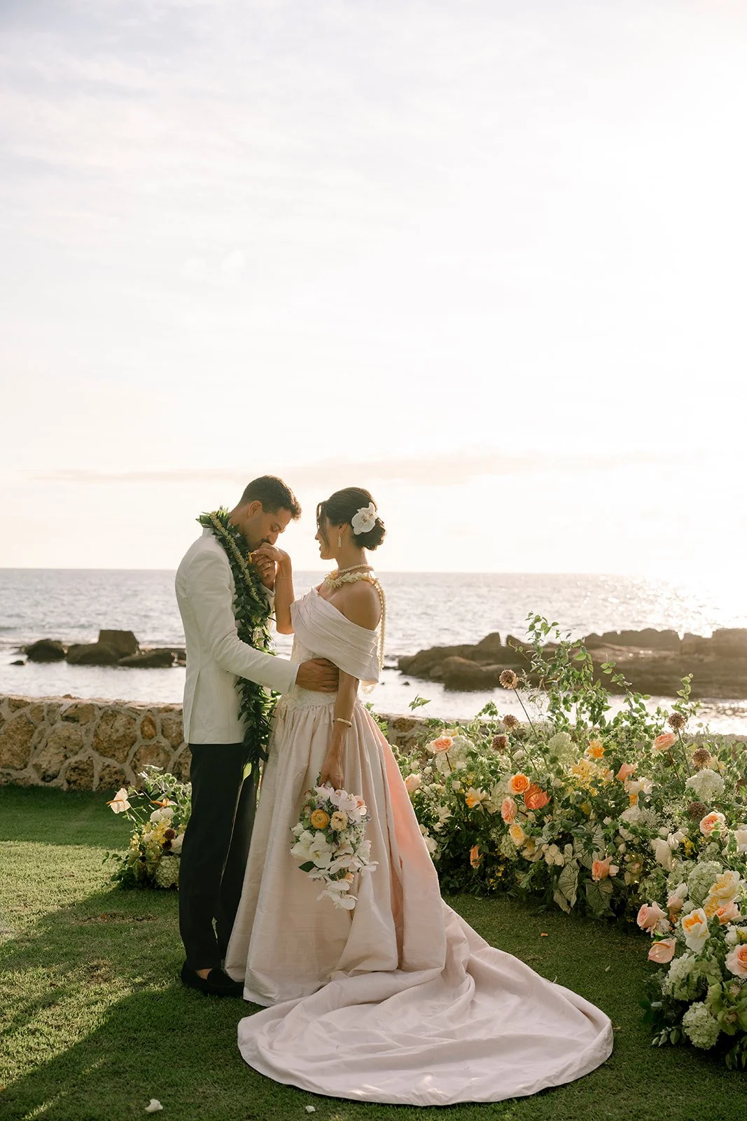 groom kissing the brides hand in front of flowers and ocean