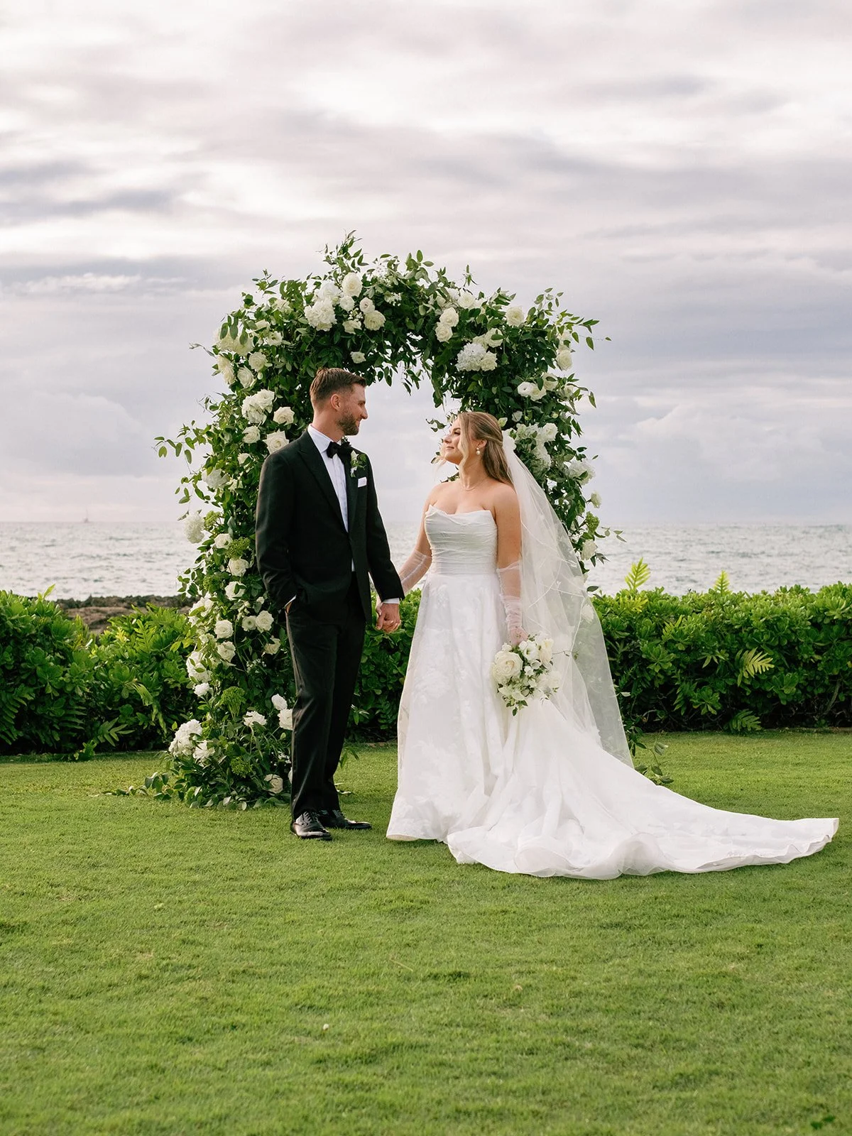 Bride and groom pose in front of their full floral arch at their ocean front Hawaii wedding.
