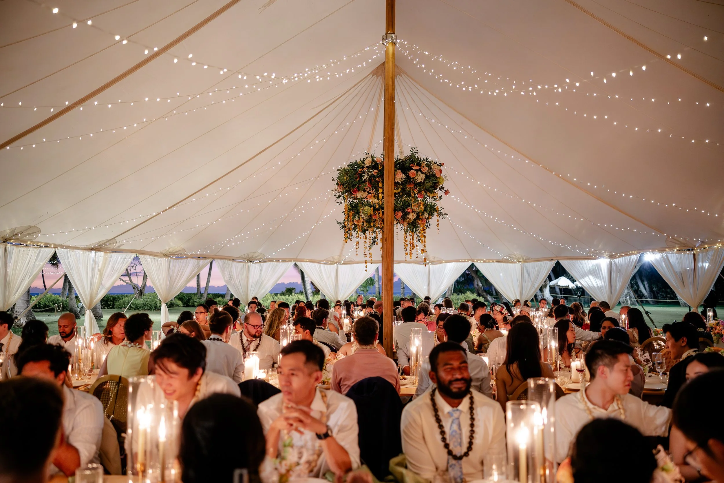 a decorated dinner reception under a large, illuminated white tent, featuring rectangle tables with floral centerpieces, and flower chandeliers