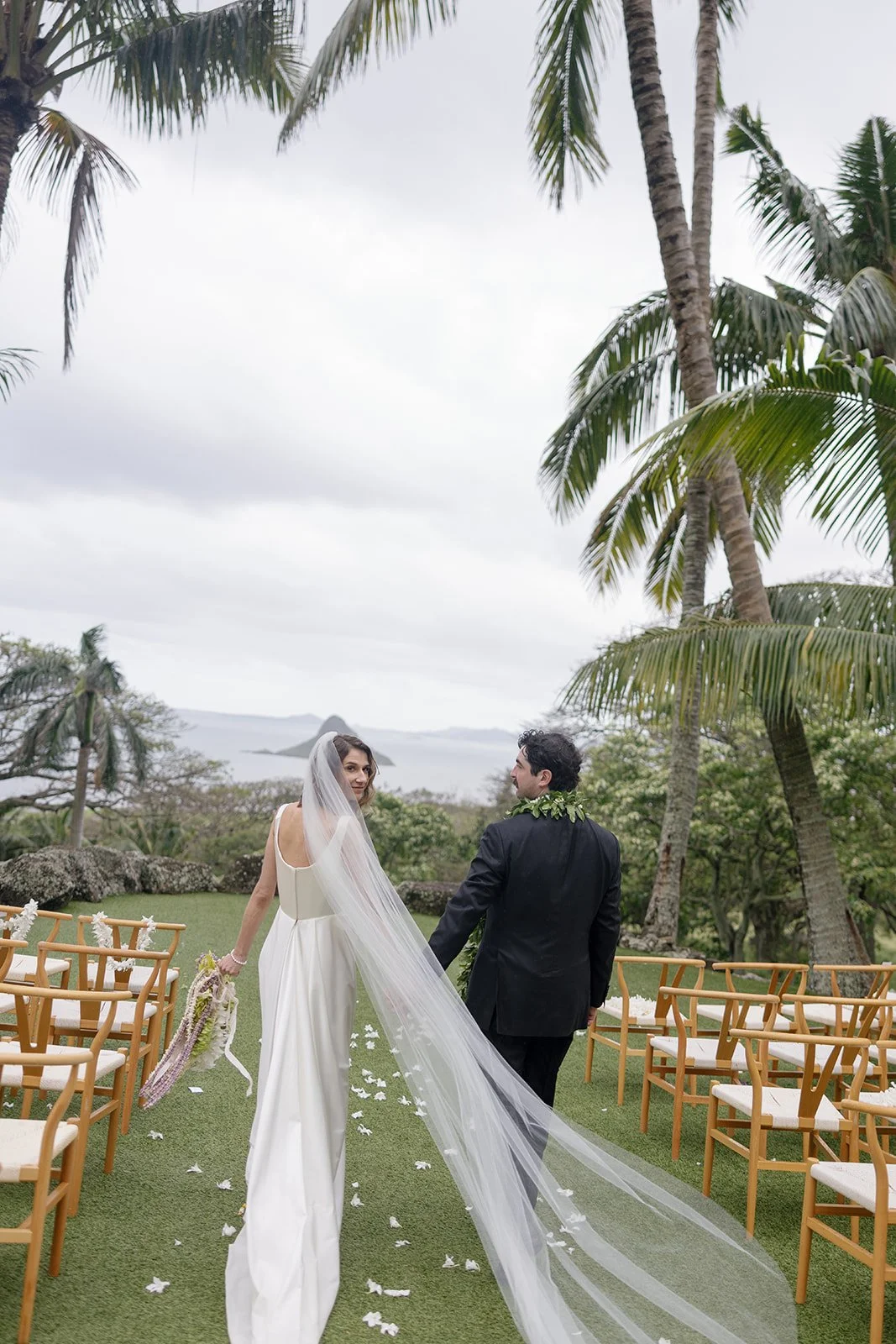Bride and groom walking hand in hand at Paliku Gardens wedding, bride looking back with cathedral veil framing her face and cascading bouquet in hand