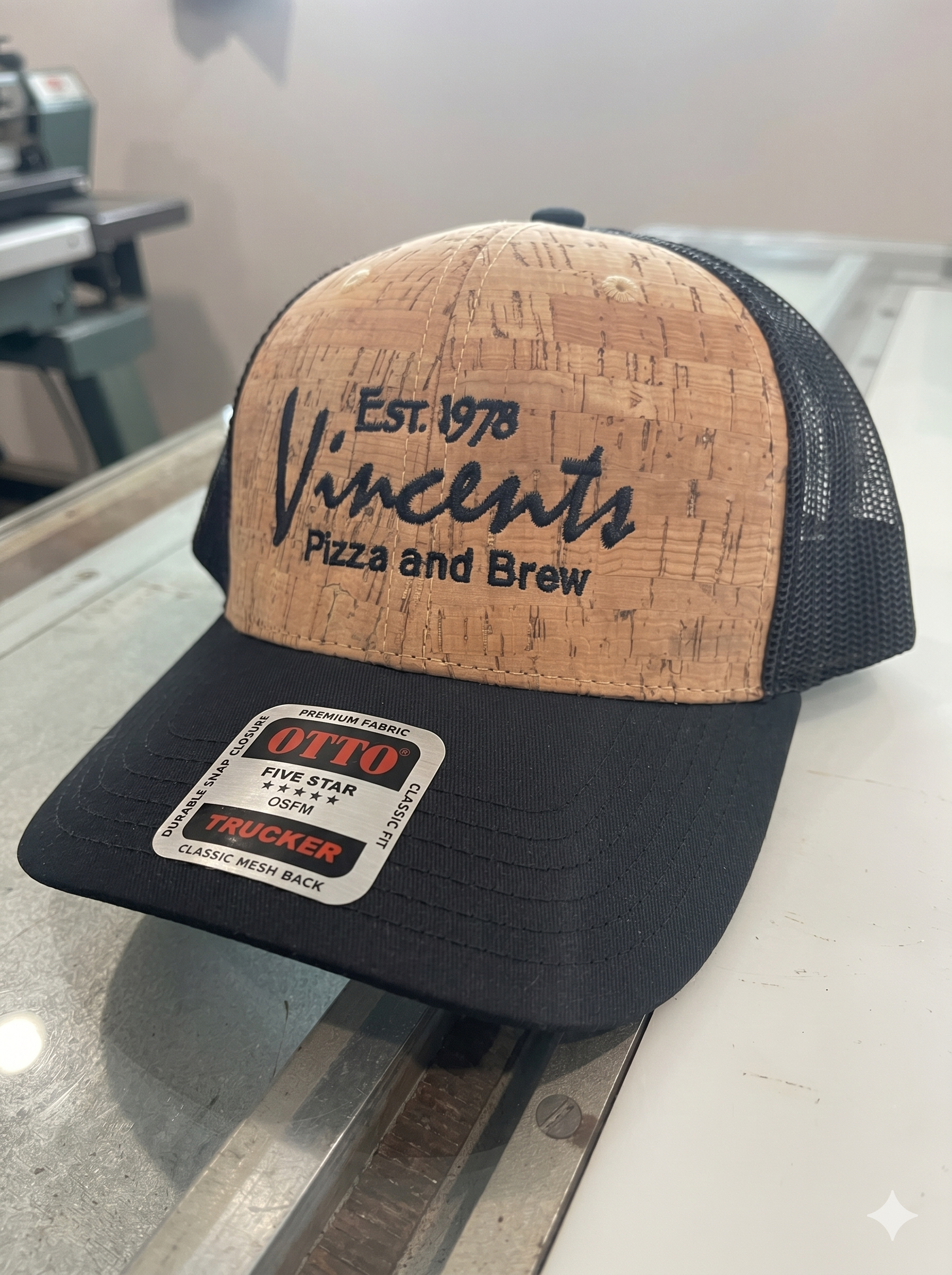 A trucker cap with a cork-style front panel that has the words "Vincent's Pizza and Brew" embroidered on it, sitting on a reflective surface.