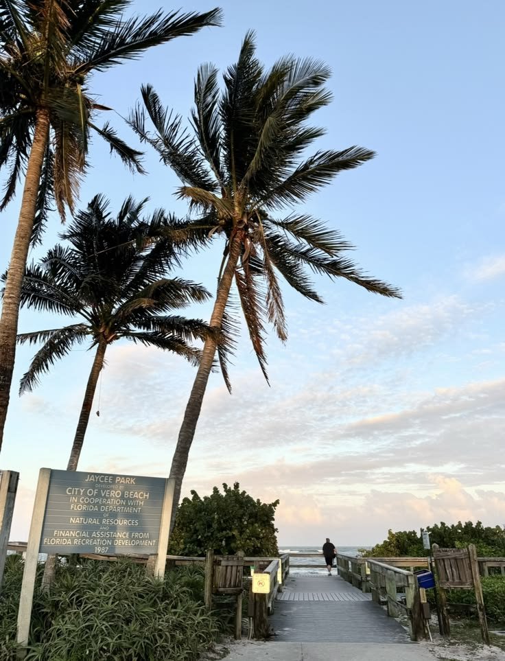 View of a boardwalk leading to the beach, with palm trees and a person walking, under a partly cloudy sky at Jaycee Park, Vero Beach, Florida.