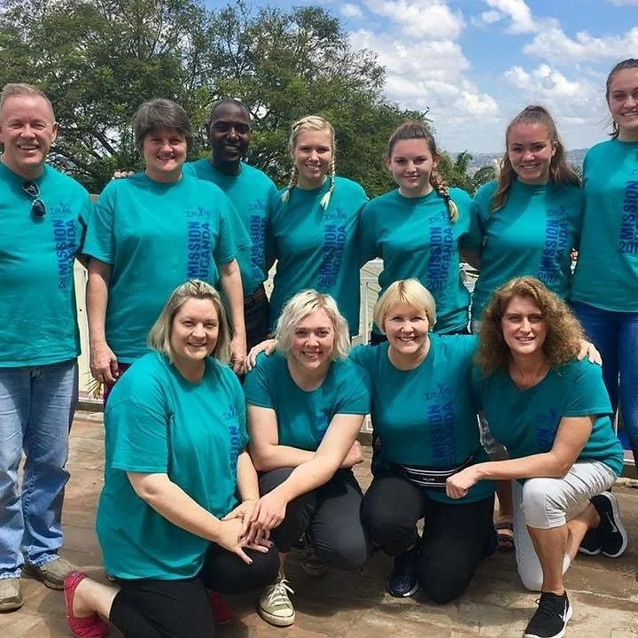 Group of ten people, seven women and three men, outdoors wearing matching turquoise T-shirts that say 'Mission Uganda 2018.'