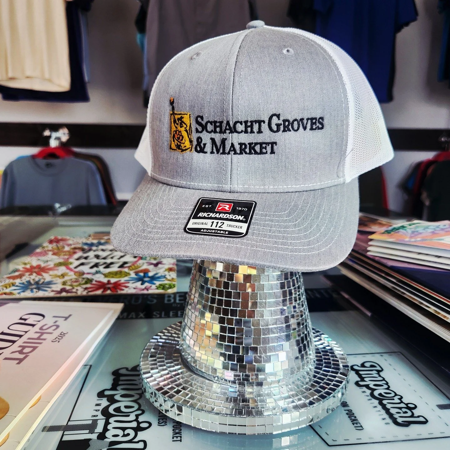 Gray baseball cap with embroidered logo and text 'Schaht Groves & Market' displayed on a shiny, mosaic-style stand in a retail store.