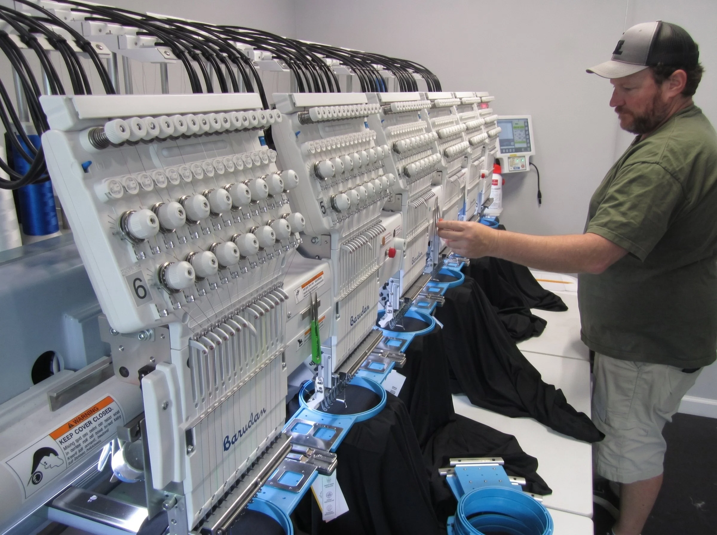 A man wearing a cap and jacket is working on embroidery machines in a brightly lit room. The machines have multiple spools of thread and various components, with some blue containers placed below them.
