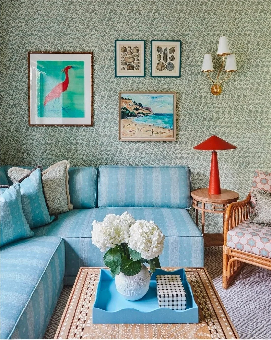 small patterned sage green wallpaper with ocean themed prints and a triple wall sconce in brass with white shades, the sofa is built in and has light blue stripes, with a cane chair and coffee table in the corner sits a small conical red table lamp