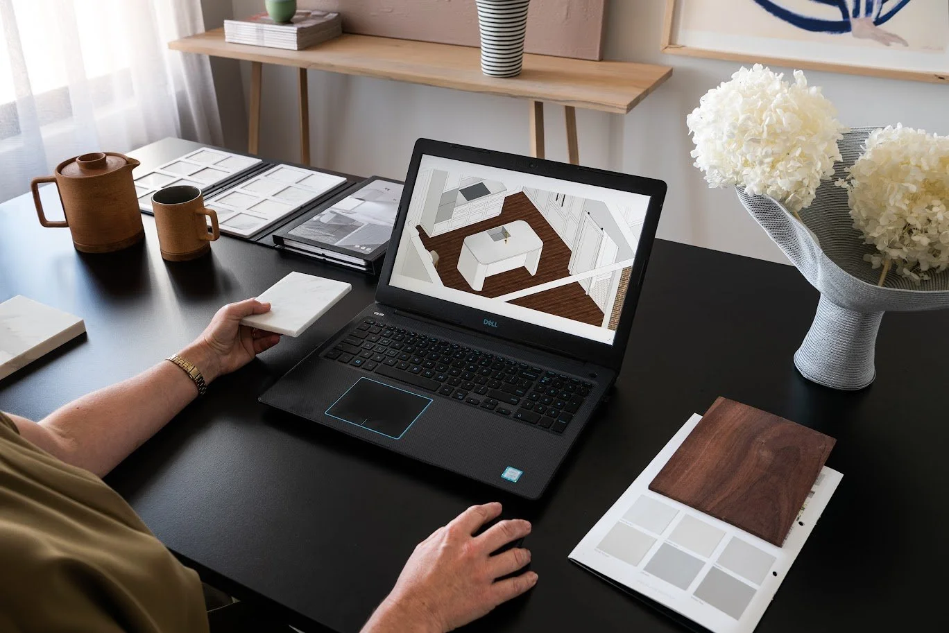 adelas desk with a stone sampler folder opened, terracotta teapot and mug, dried white hydrangeas in a vase and a drawing of a kitchen on her laptop