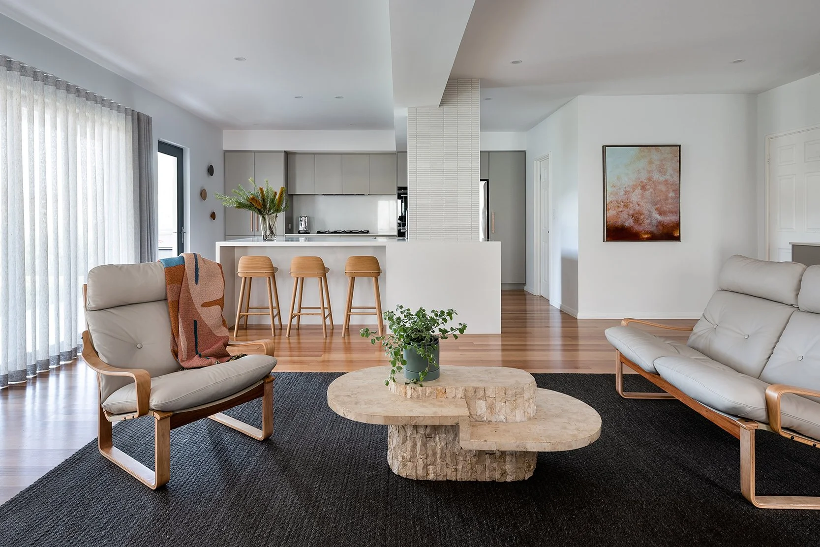an open plan kitchen and living room with honey coloured timber floors, large sliding doors to the garden with linen sheer curtains.  The restored leather chairs and vintage limestone coffee table are grounded by a dark grey wool flatweave rug